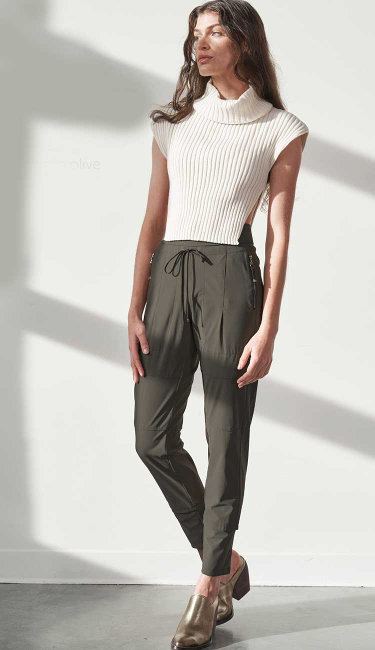 Woman wearing a white turtleneck and olive drawstring pants against a plain wall.