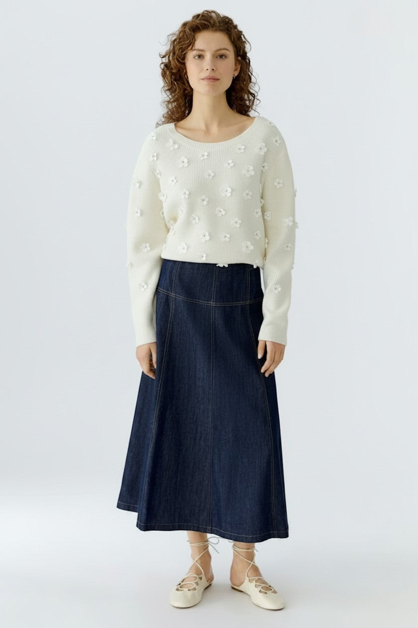 Woman wearing a white sweater with floral patterns and a long navy skirt on a white background.