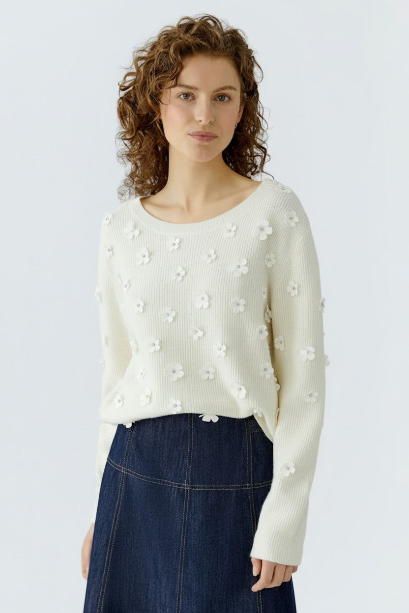 Woman wearing a white sweater with floral embellishments and a blue skirt on a light gray background.