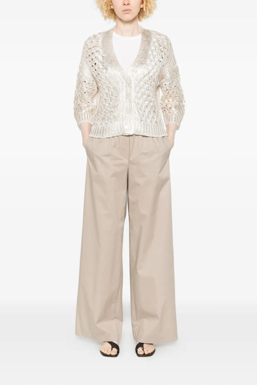 Person wearing a gold lace cardigan and beige pants on a white background.