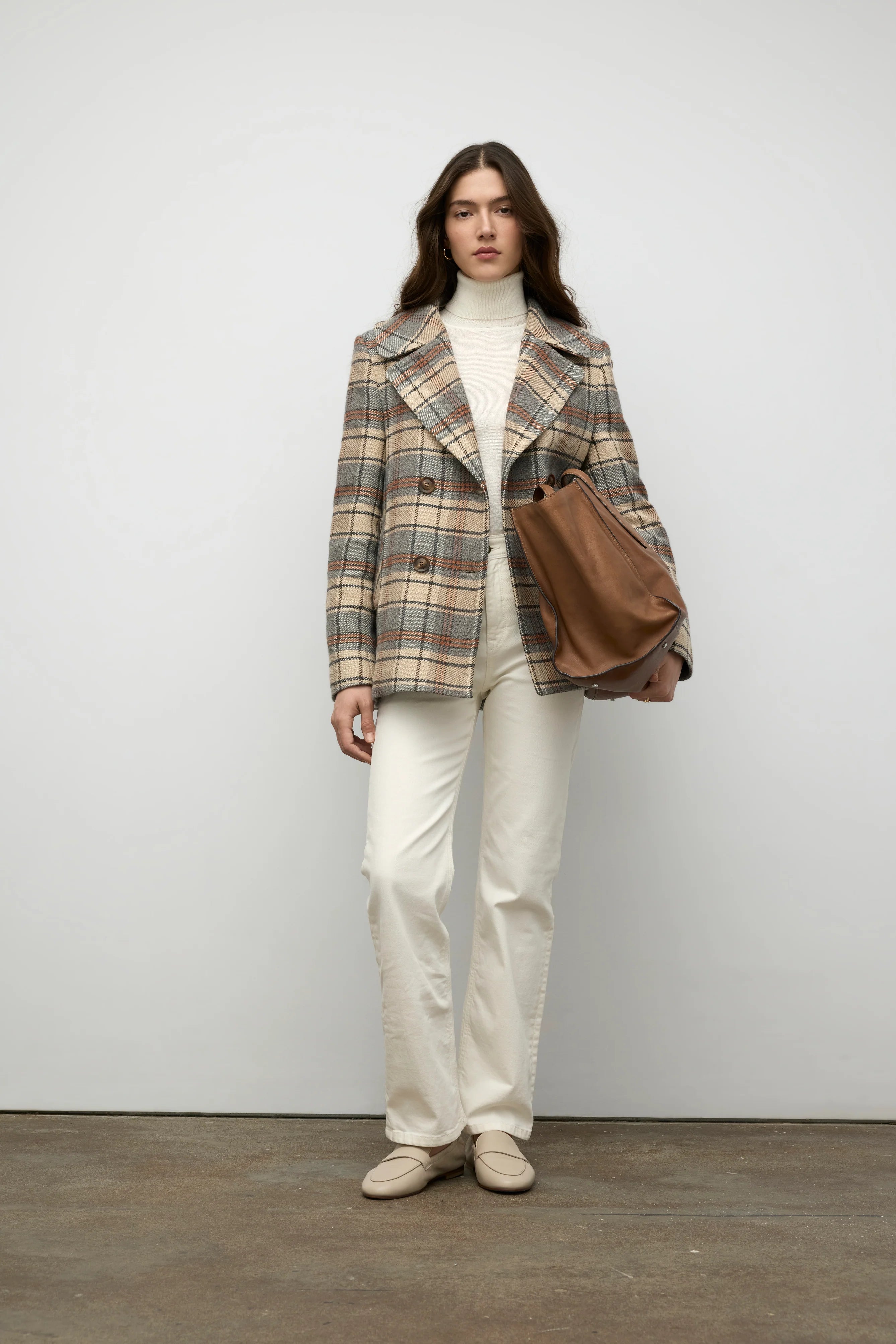 Woman wearing a plaid coat, white pants, and holding a brown leather bag against a plain background.