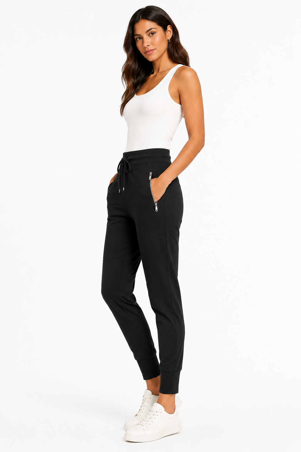 A woman modeling black jogger-style pants with a drawstring waist and zippered side pockets, paired with a white tank top and white sneakers, against a plain white studio background.