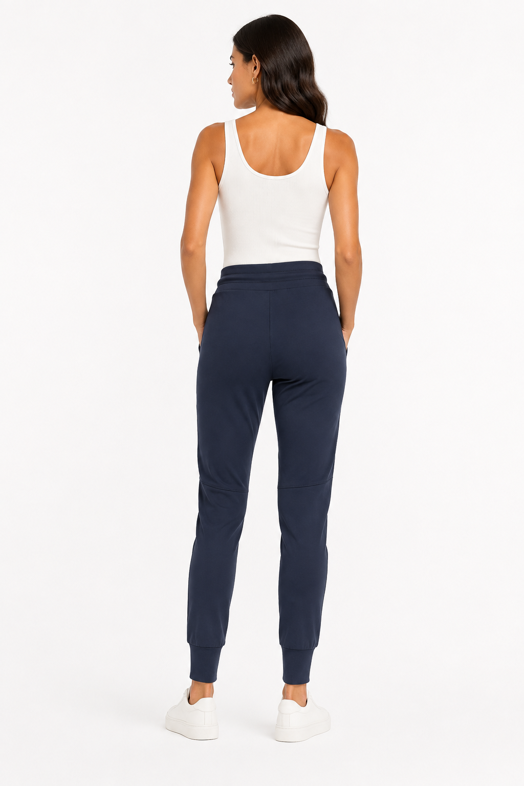 A woman shown from the back wearing navy blue jogger sweatpants, a white tank top, and white sneakers against a plain white studio background.