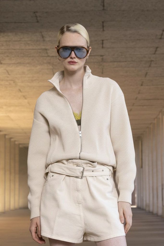 Woman wearing sunglasses and a beige outfit in a long, narrow hallway.