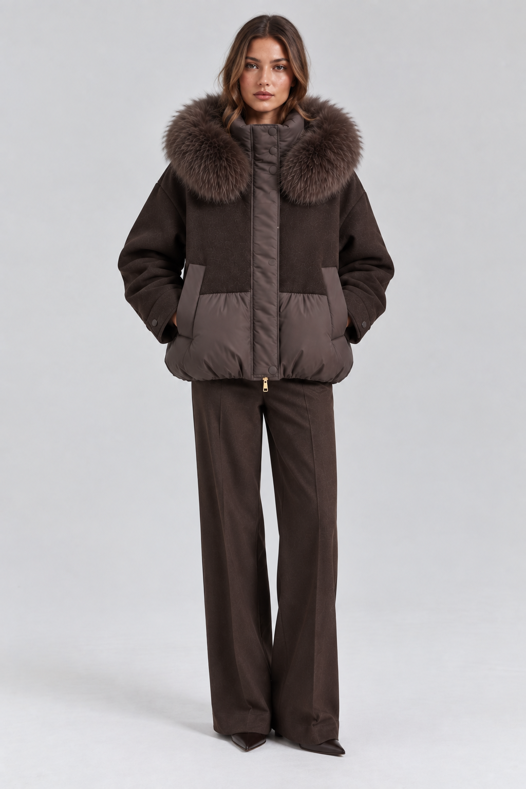 Model wearing a dark brown mixed-media puffer jacket with a plush fur-trimmed hood, wool-textured upper panels, padded lower section and gold zipper detail, styled with wide-leg brown trousers.