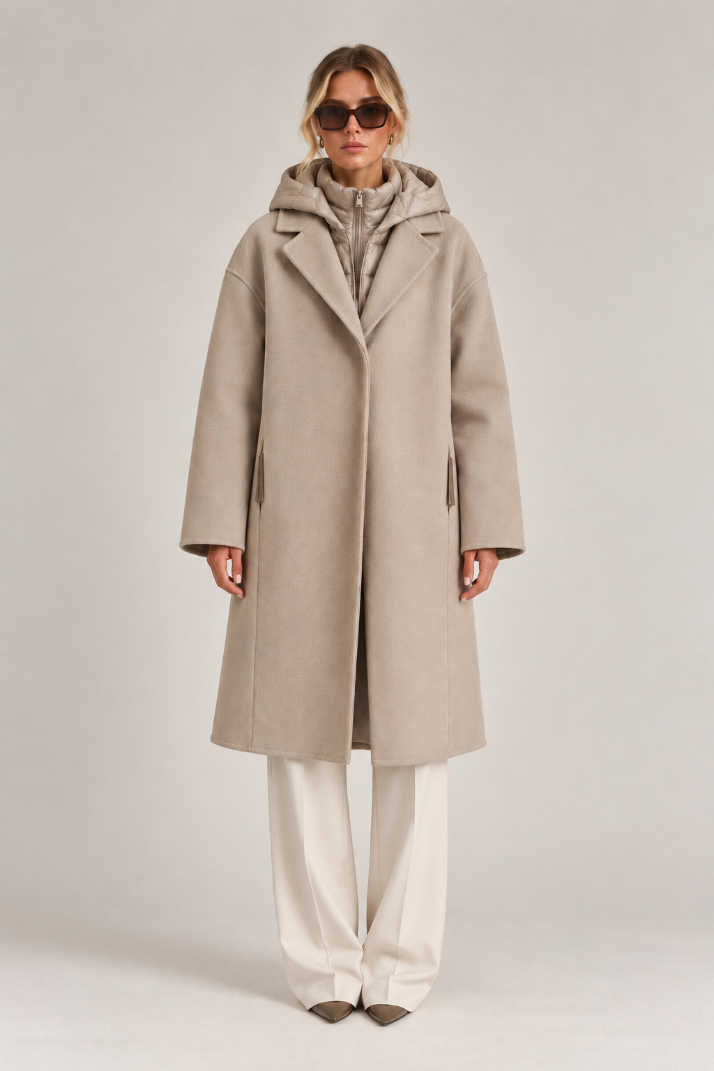 Front view of model wearing a taupe long coat with quilted hooded insert, styled with ivory wide-leg trousers.