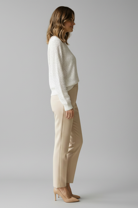 Woman wearing a white sweater with diamond detailing and beige pants against a beige background.