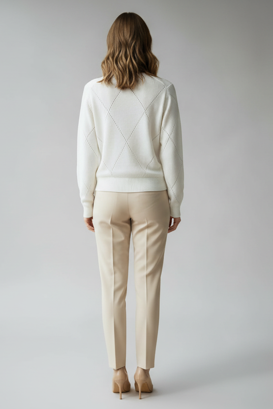 The back of a woman wearing a white sweater with diamond detailing and beige pants against a plain background.
