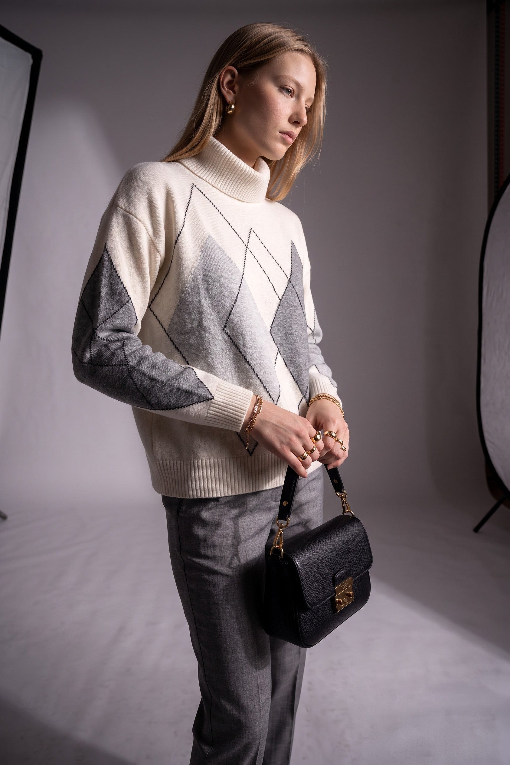 A woman modeling a cream and gray geometric knit turtleneck sweater, paired with gray trousers and carrying a black leather shoulder bag with gold hardware in a studio setting.
