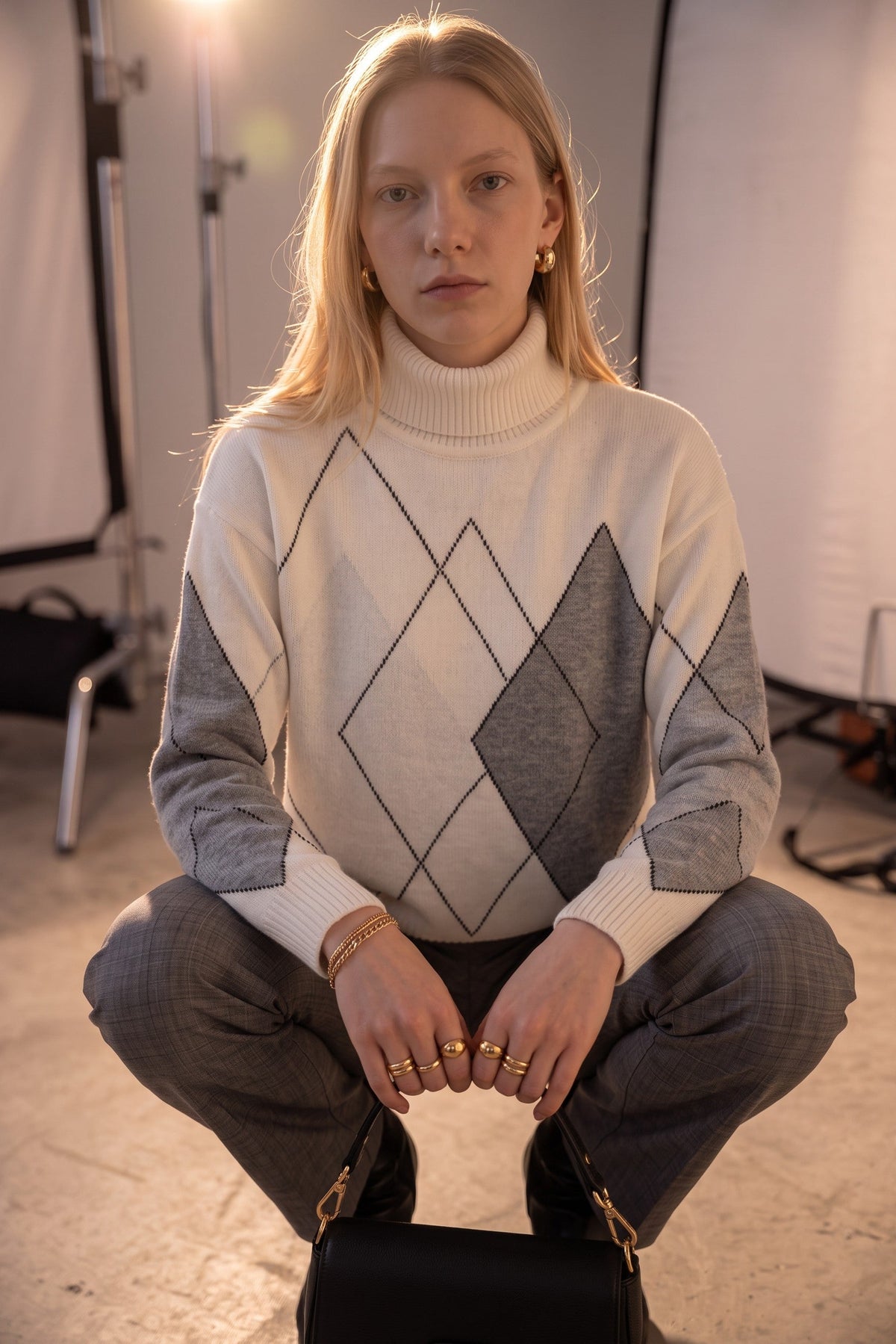 A model crouching in a studio setting, wearing a cream and gray argyle knit turtleneck sweater, gray plaid trousers, and gold jewelry.