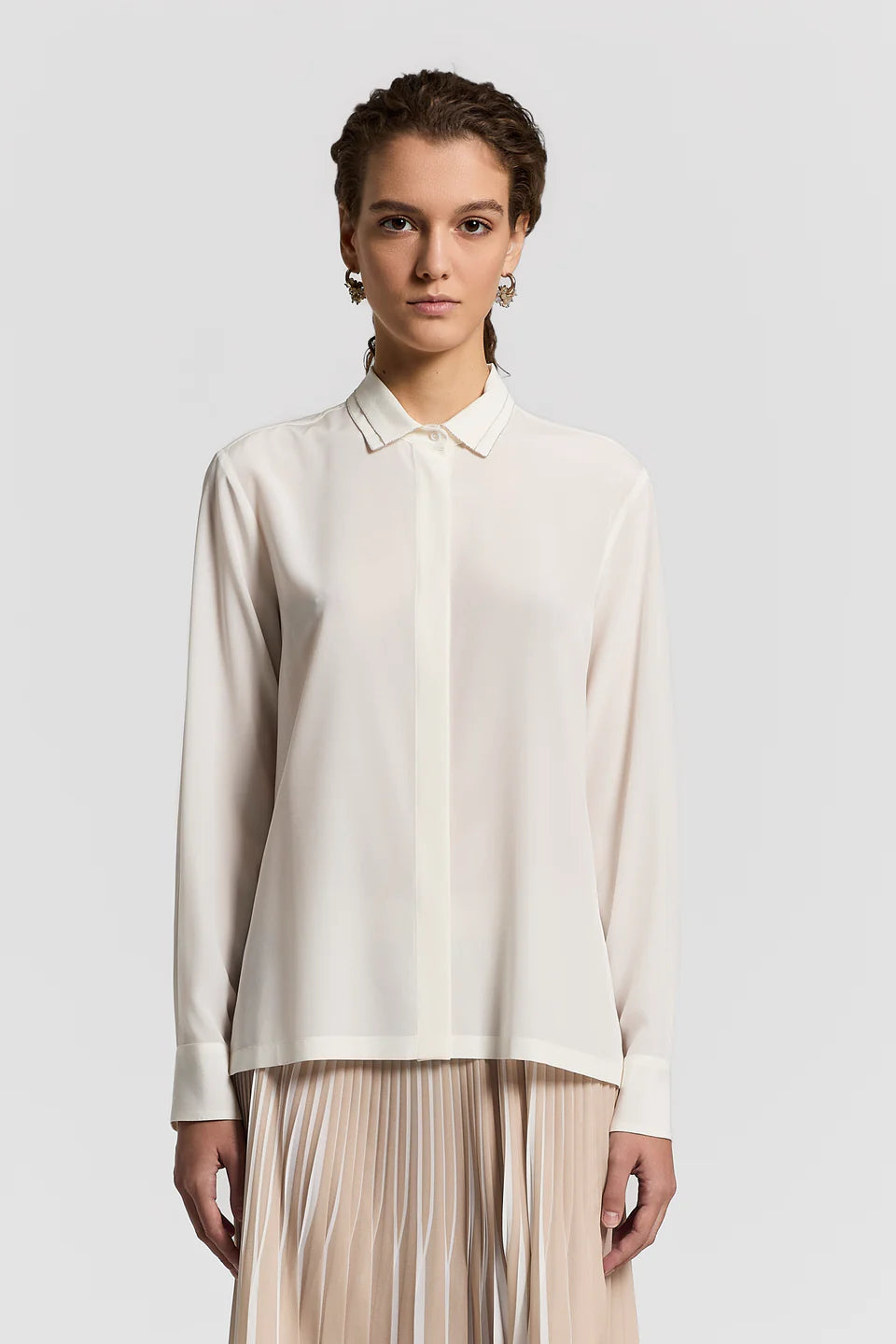 Woman wearing a white blouse and beige pleated skirt on a light gray background.