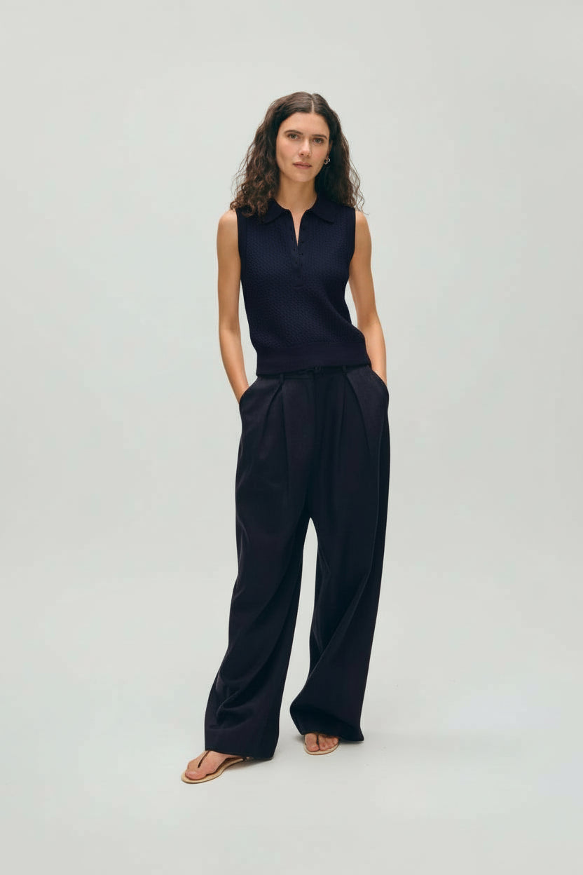 Woman wearing a navy vest with navy trousers and sandals on a gray background.