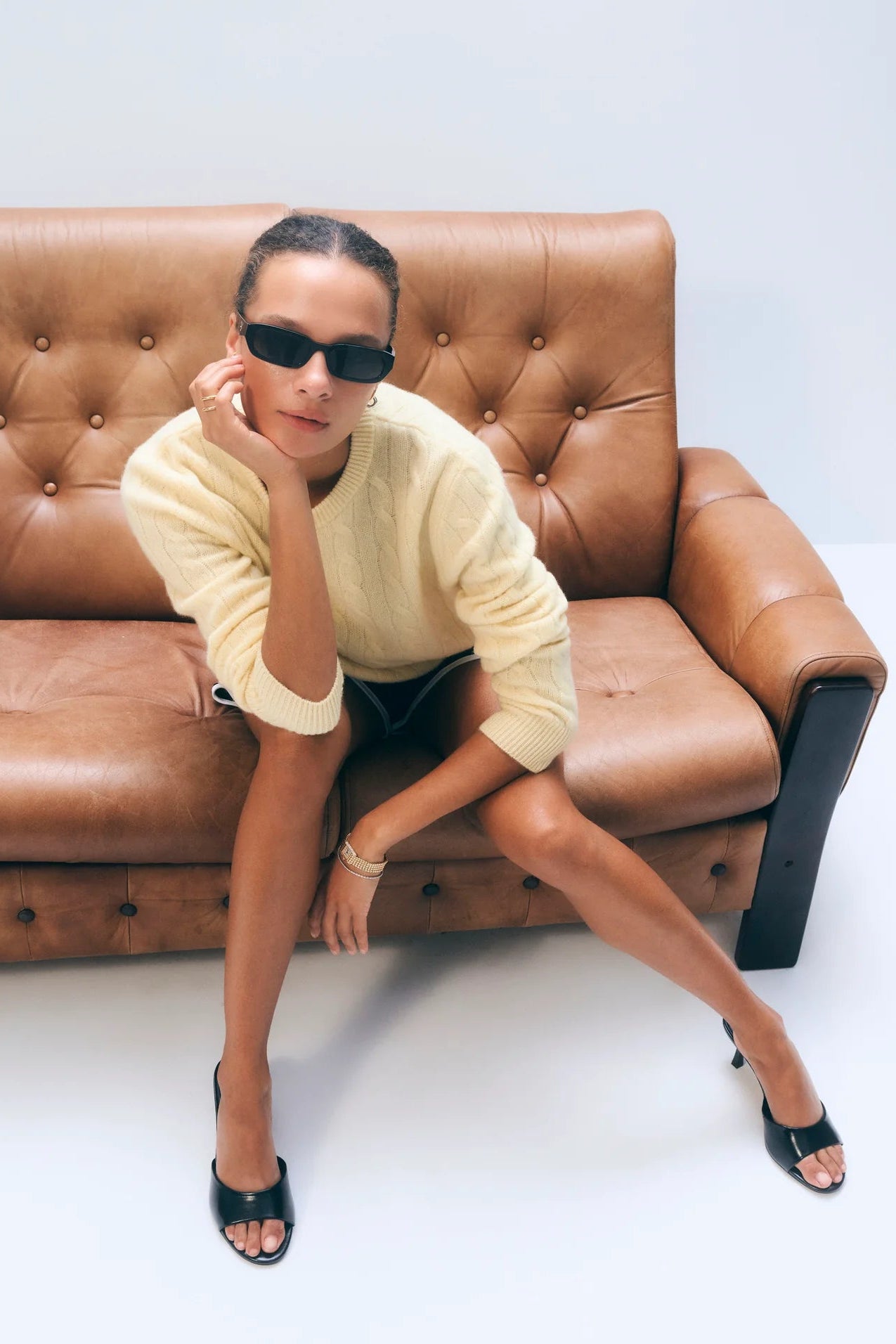 Woman sitting on a brown leather sofa wearing sunglasses and a yellow sweater.