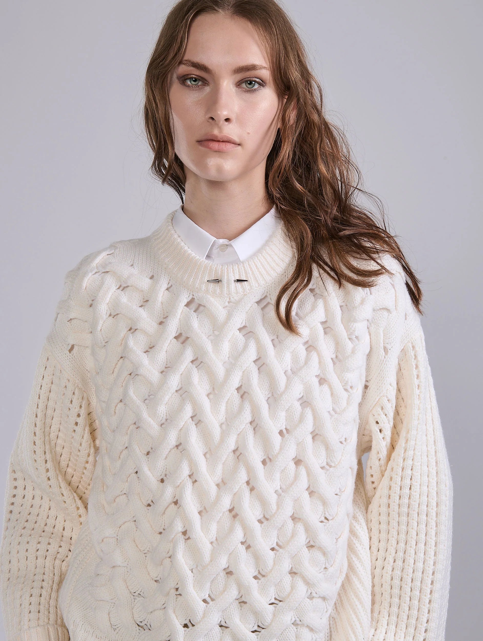Woman wearing a white cable knit sweater over a white shirt against a plain background.