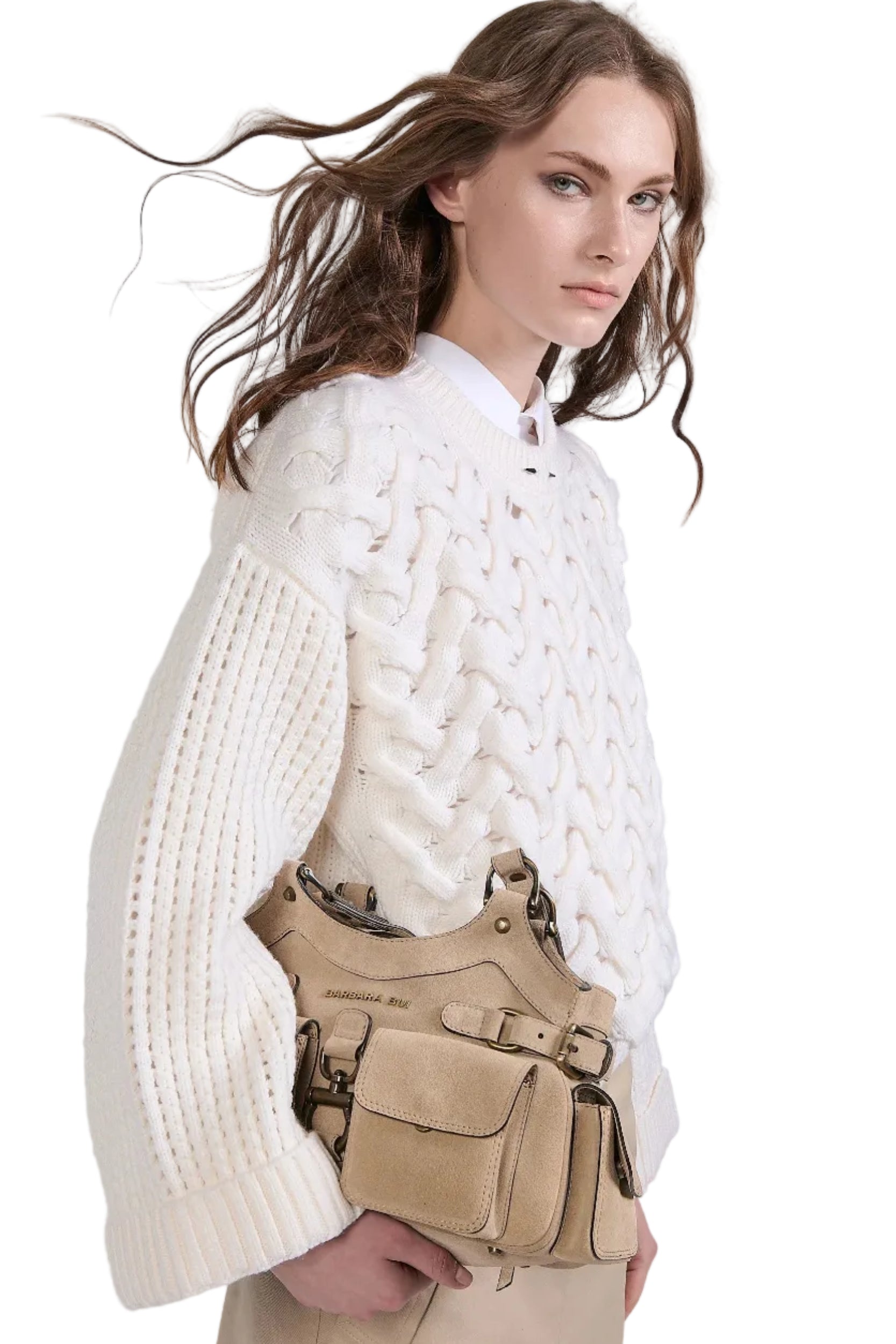 Woman wearing a white cable knit sweater and beige pants with a beige bag on a light gray background.