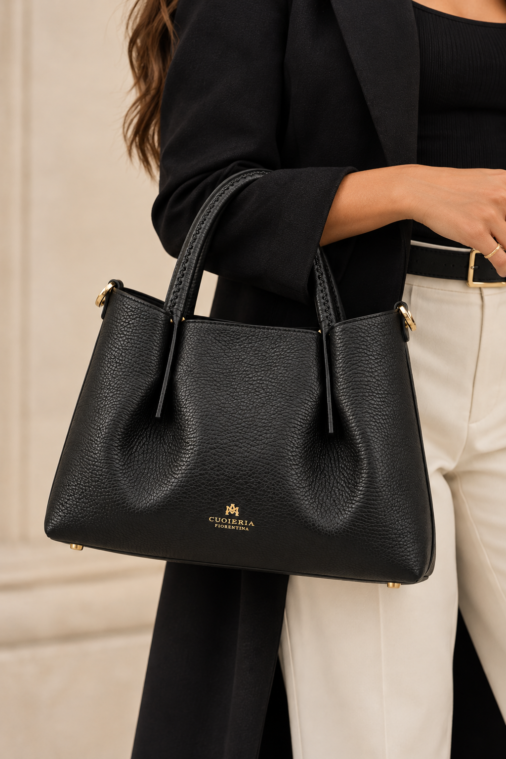 Close-up model view of the Cuoieria Fiorentina Candy Medium Tote in black pebbled calfskin leather, featuring curved stitched handles, gold-tone hardware, and signature logo detailing, styled with a black blazer and cream trousers.
