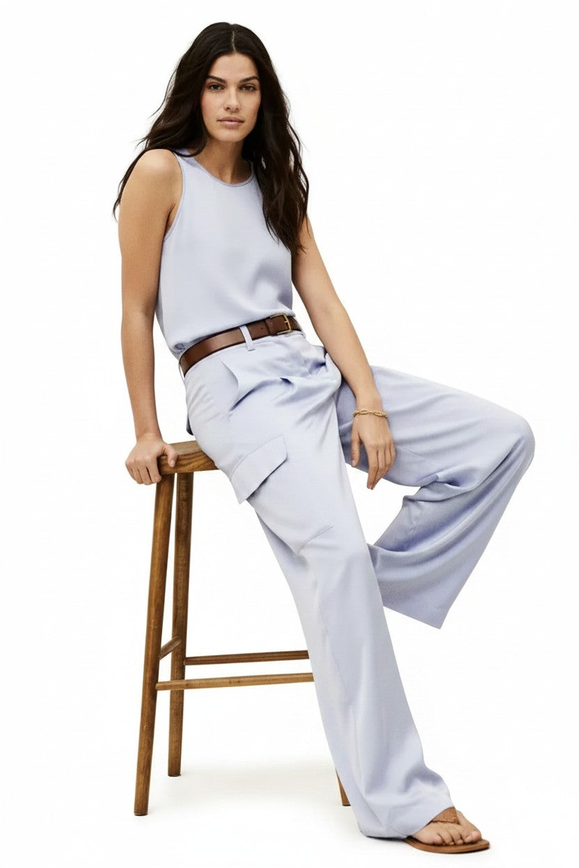 Woman sitting on a wooden stool wearing a lavender sleeveless top and wide-leg pants against a white background.