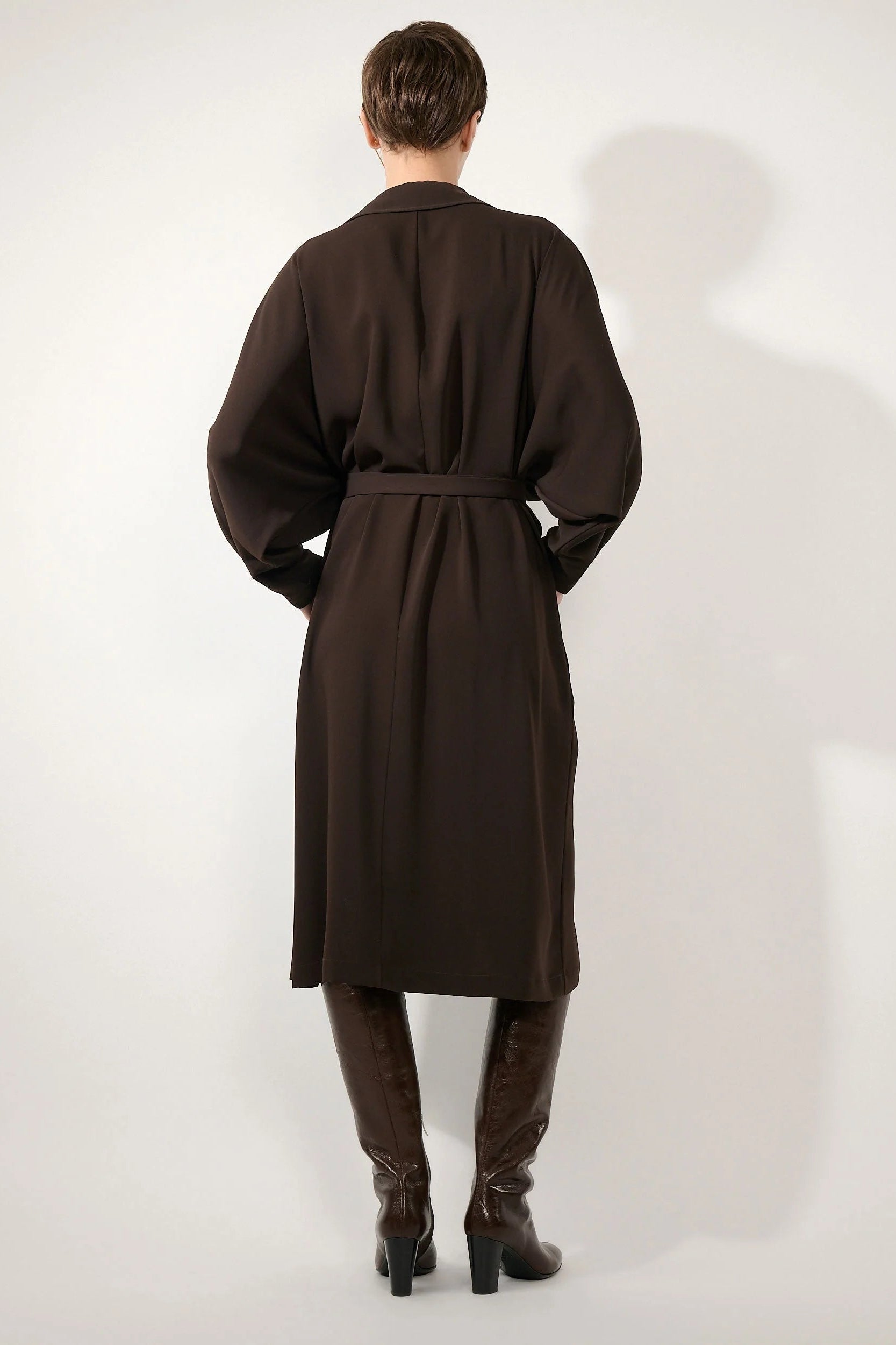 A model viewed from the back wearing a dark brown belted coat or dress over a matching dark brown top and brown knee-high boots, standing against a plain light gray studio background.