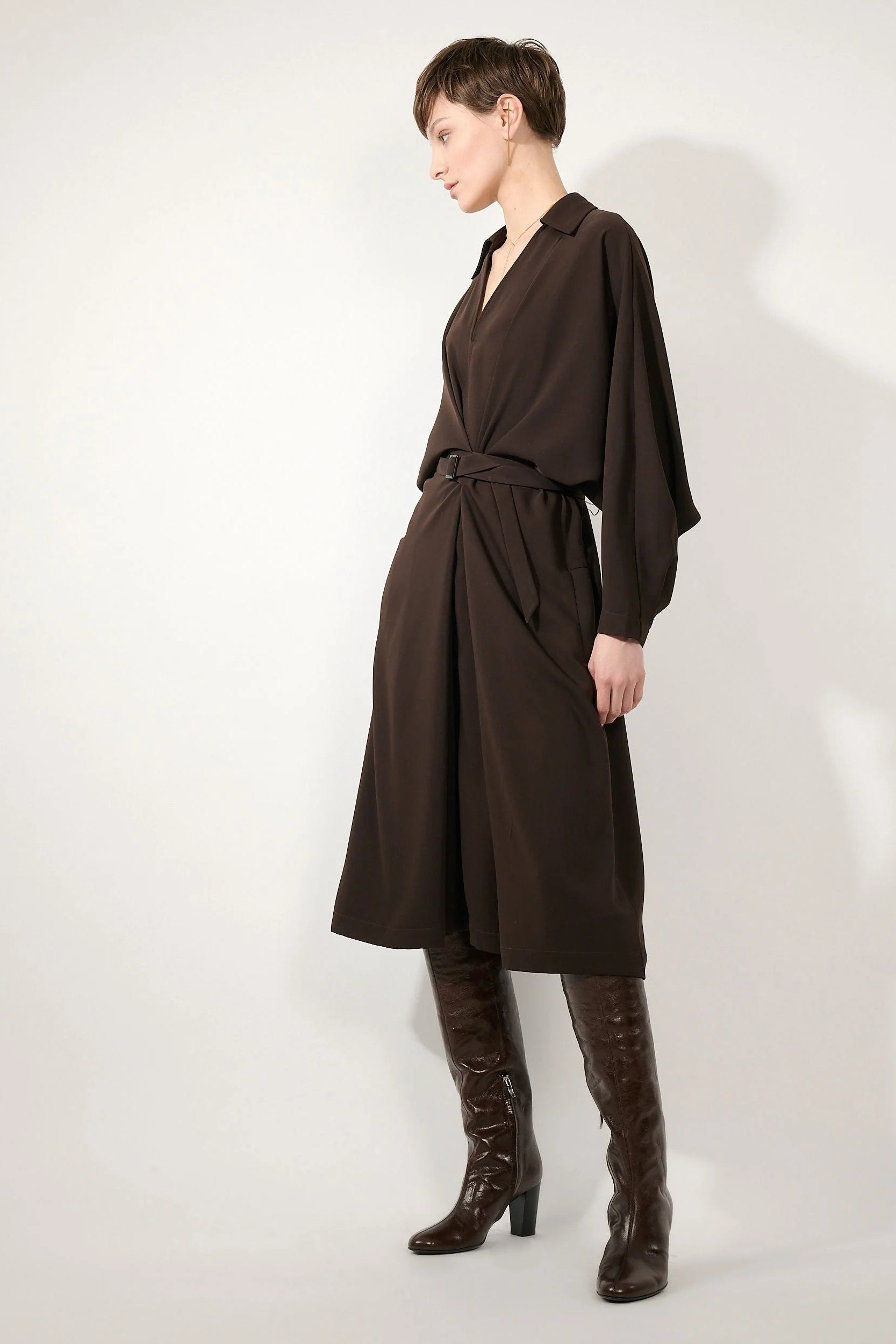 A model wearing a dark brown wrap dress with a tie waist and matching long sleeves, paired with dark brown knee-high boots, photographed against a light gray studio background.