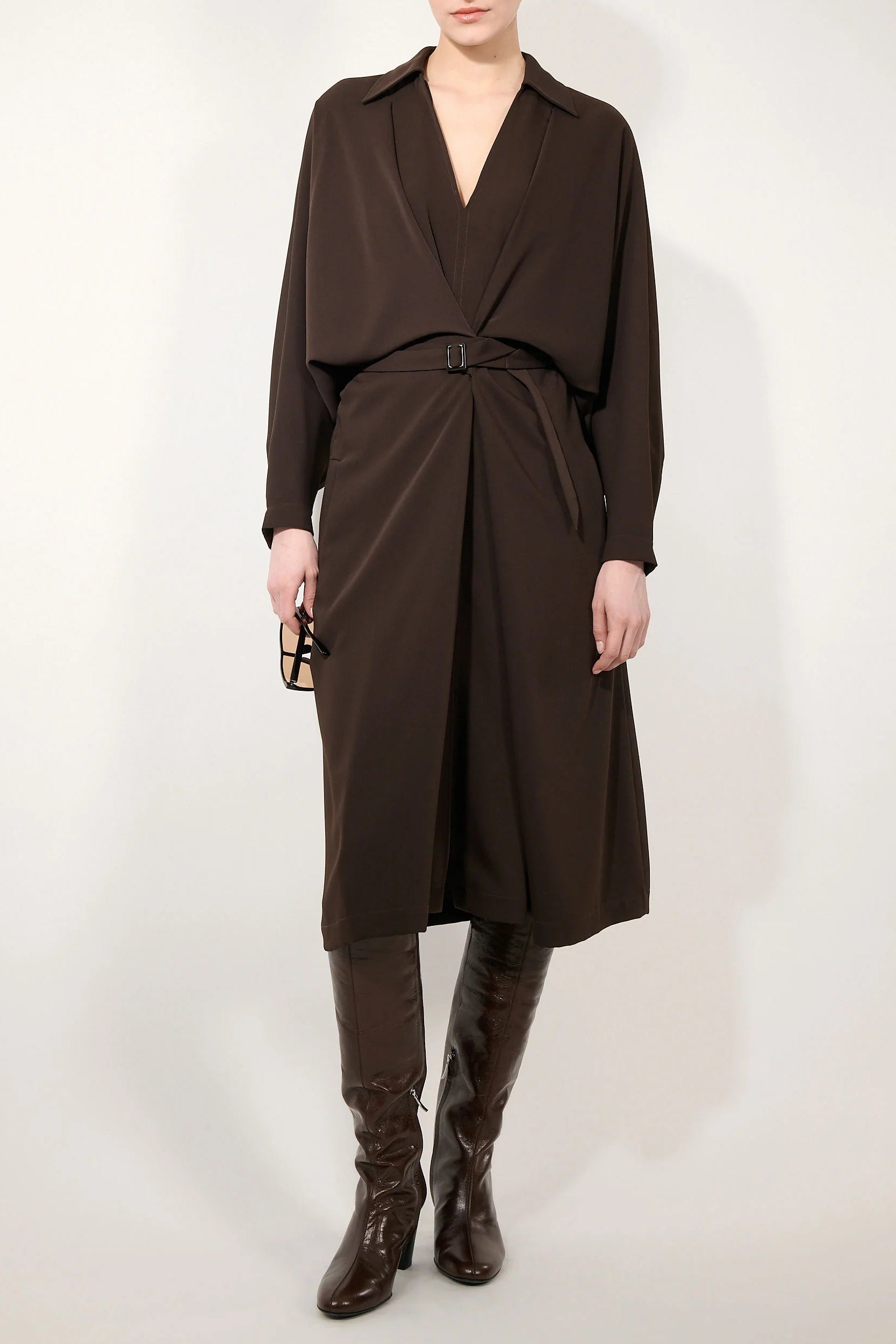 A model wearing a dark brown wrap-style midi dress with a matching waist tie belt, paired with dark brown knee-high leather boots, against a plain off-white studio background.