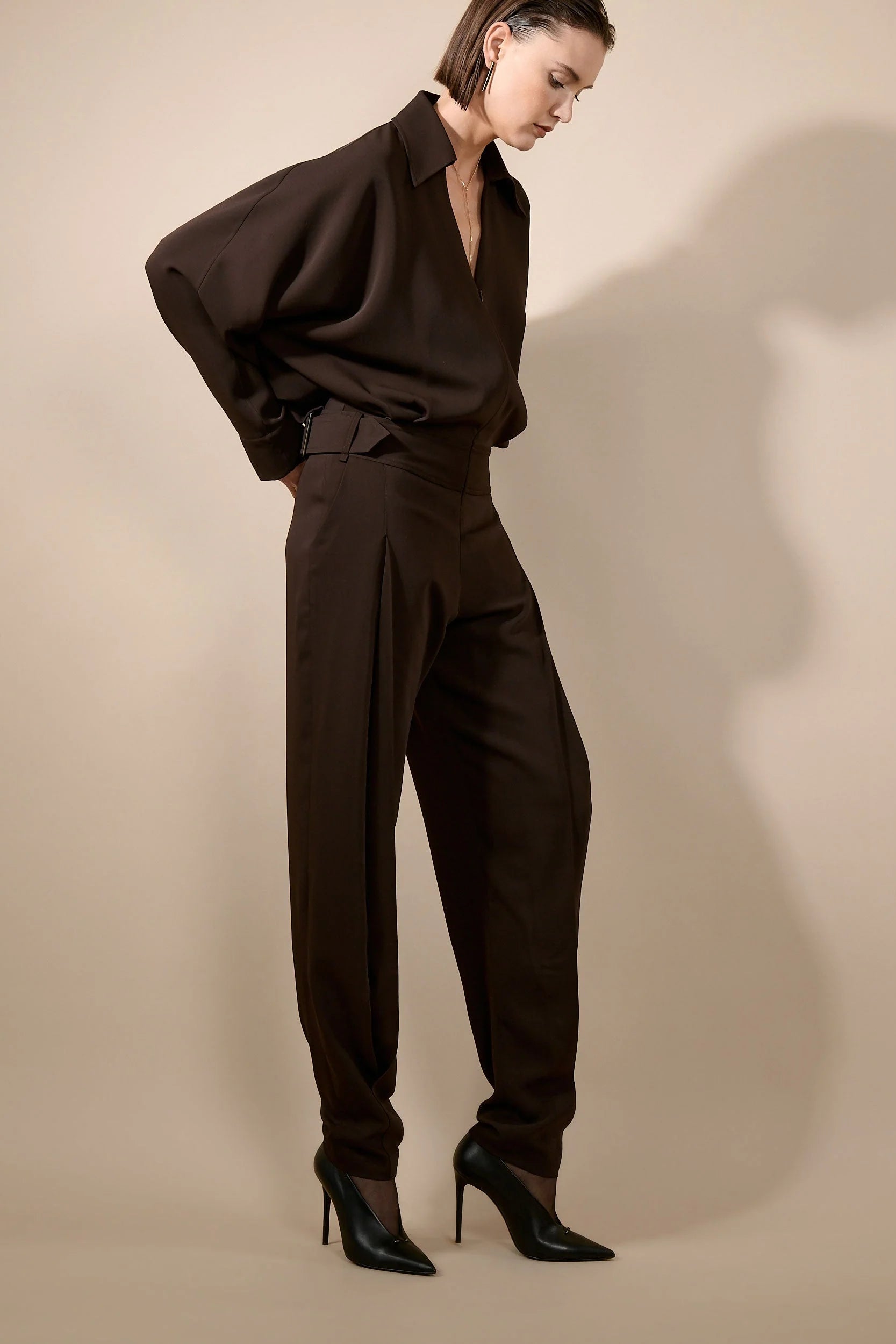 A model wearing a dark brown two-piece set consisting of a loose, oversized long-sleeve collared top and matching high-waisted pleated trousers, paired with black pointed-toe heels, against a warm beige studio background.