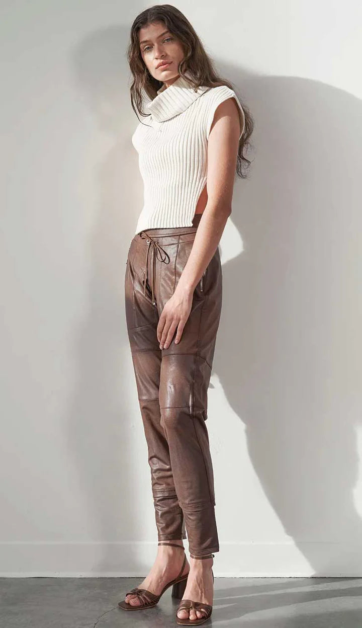 Woman wearing a white sleeveless top and brown leather pants against a white wall.