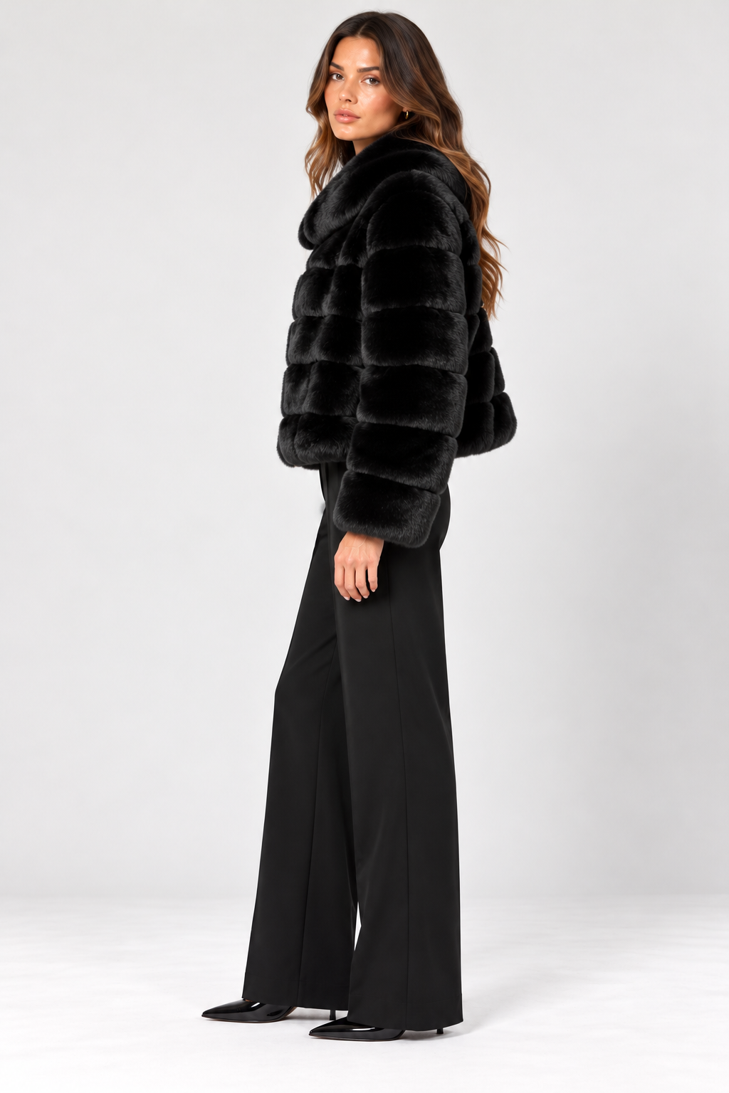 Side view of model wearing the Suprema Black Rex Rabbit Fur Bomber Jacket, featuring luxurious genuine Rex rabbit fur, horizontal fur panels, an oversized fold-down collar, long sleeves, and a relaxed hip-length silhouette. Styled with tailored black wide-leg trousers and pointed-toe heels against a neutral studio background.