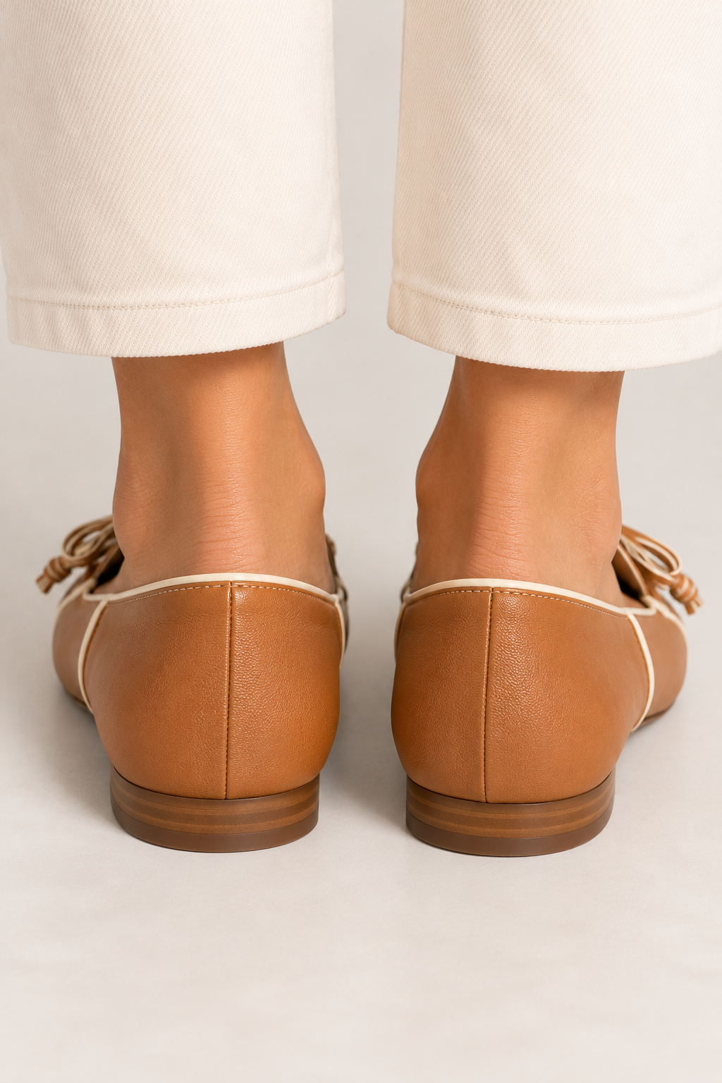 Back-view close-up of model wearing RODO Antilope camel leather loafers featuring smooth leather heel counters, ivory contrast piping, delicate stitching, and low stacked heels, styled with cropped cream trousers.