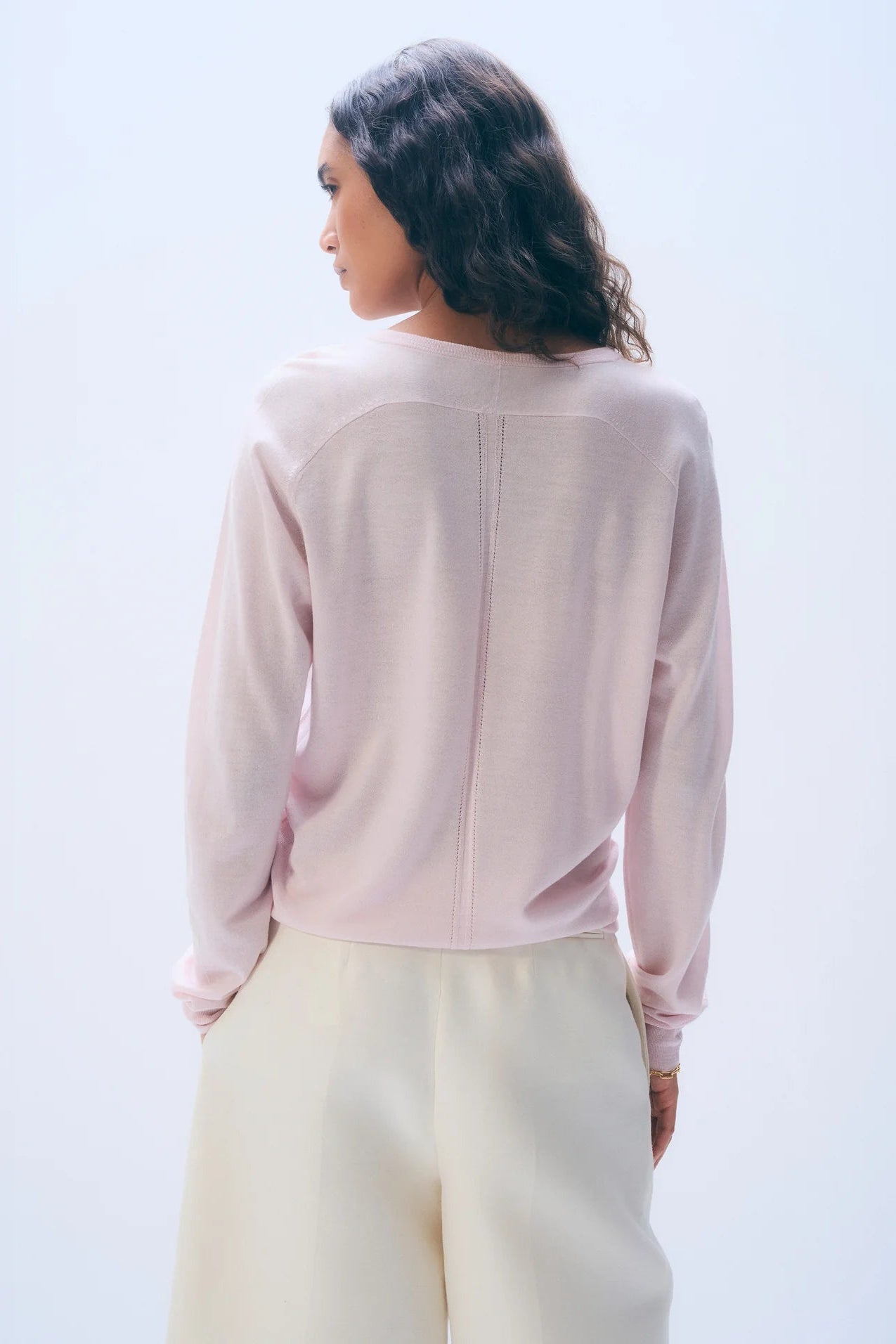 The back of a woman wearing a light pink long-sleeve sweater and white pants against a light grey background,