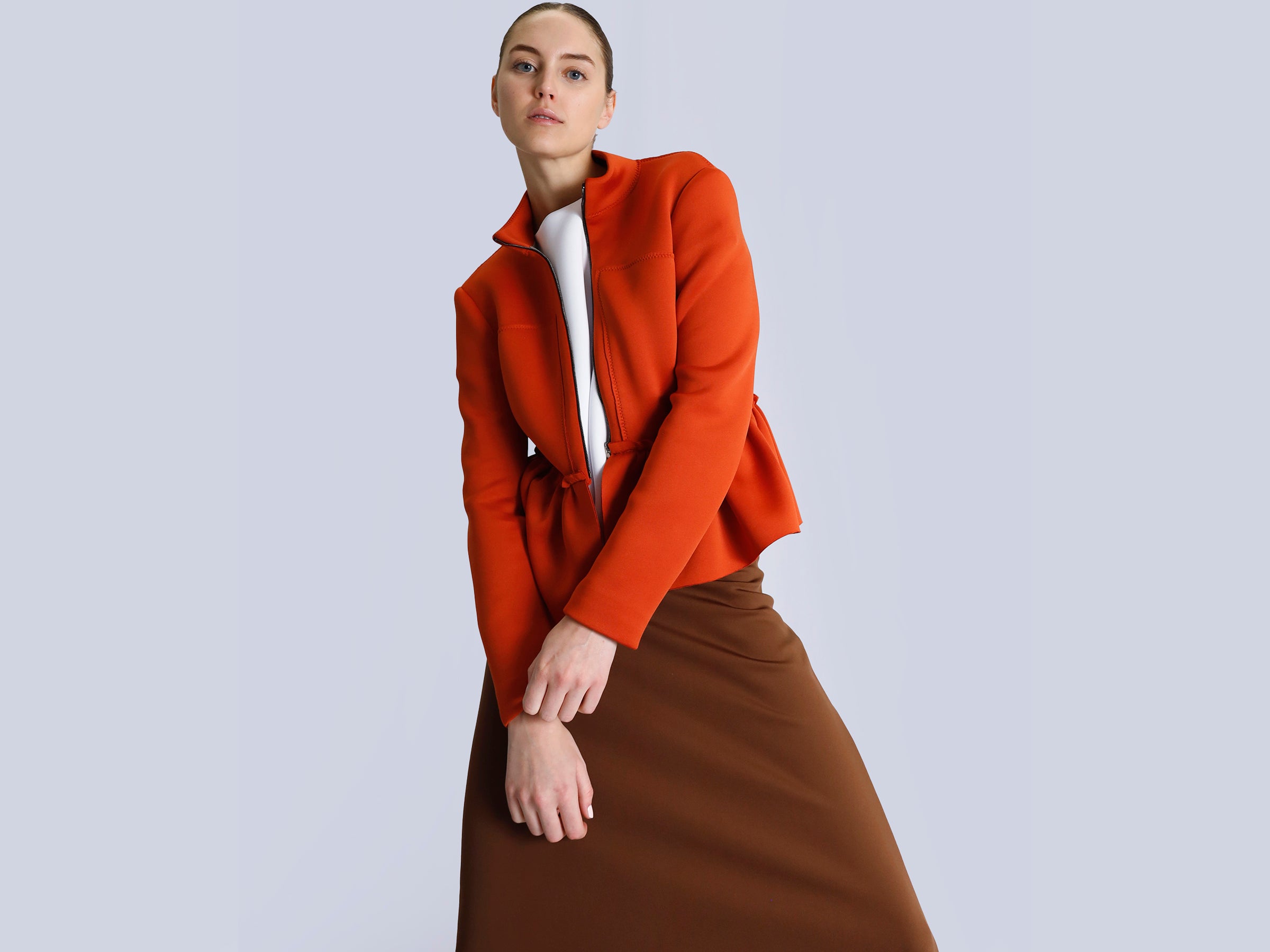 Person wearing an orange jacket and brown skirt on a light gray background