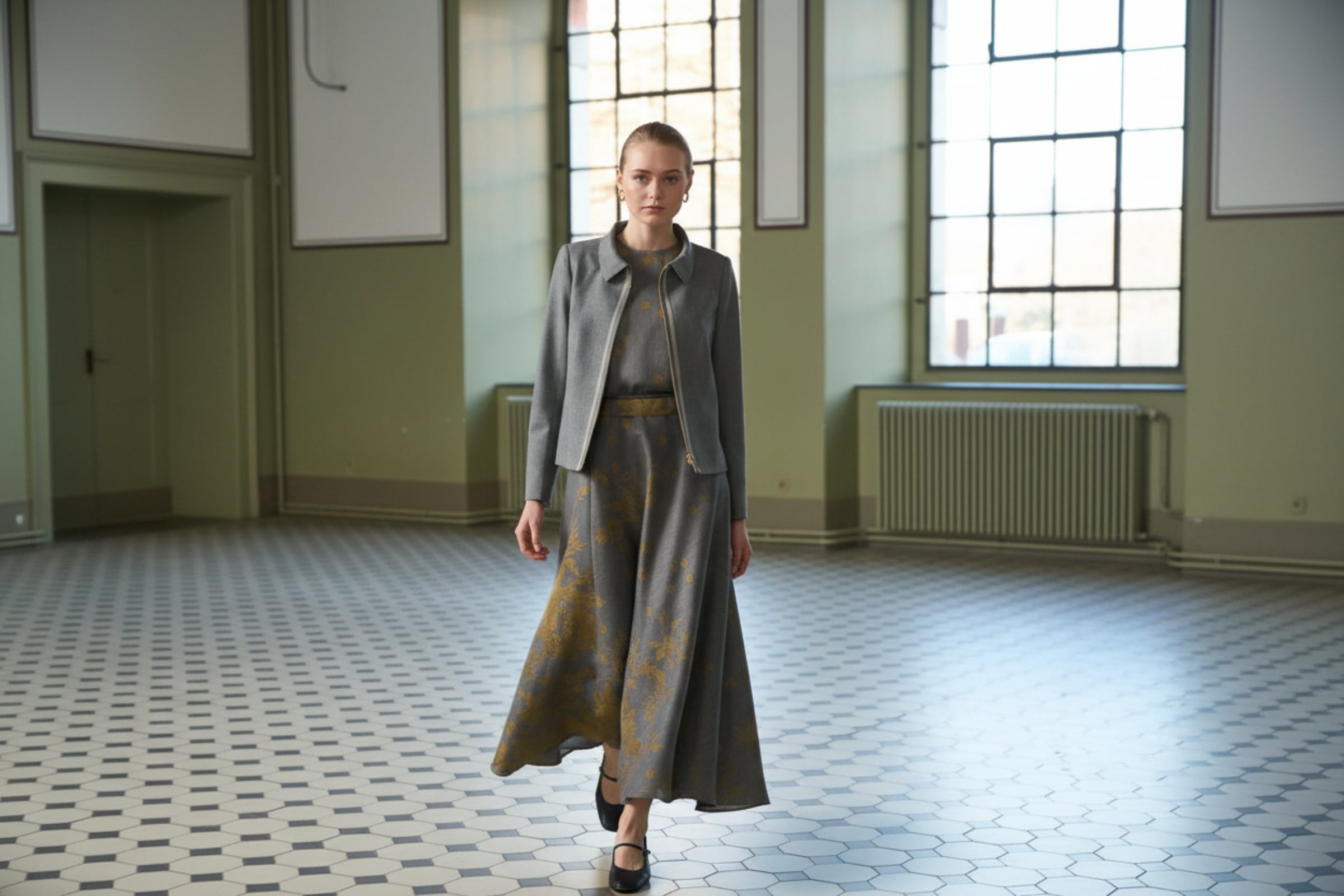 Woman in a gray outfit standing in a room with large windows.
