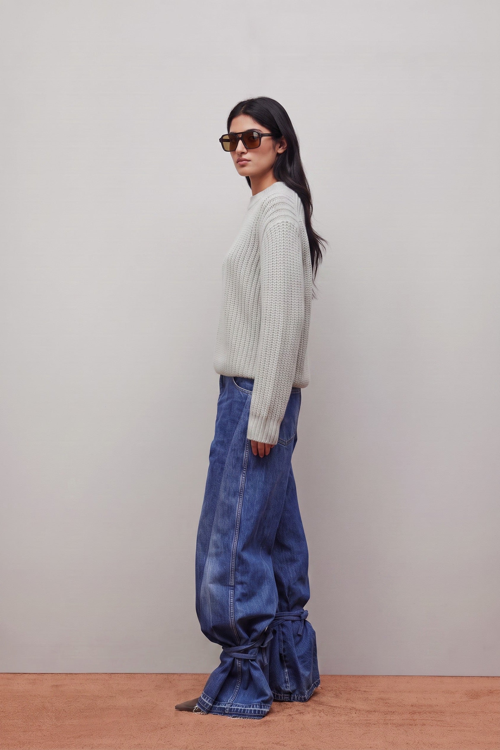 A woman stands in profile wearing a light gray ribbed knit sweater, wide-leg blue jeans with gathered ruffle hems, and dark sunglasses against a plain light gray wall and tan floor.
