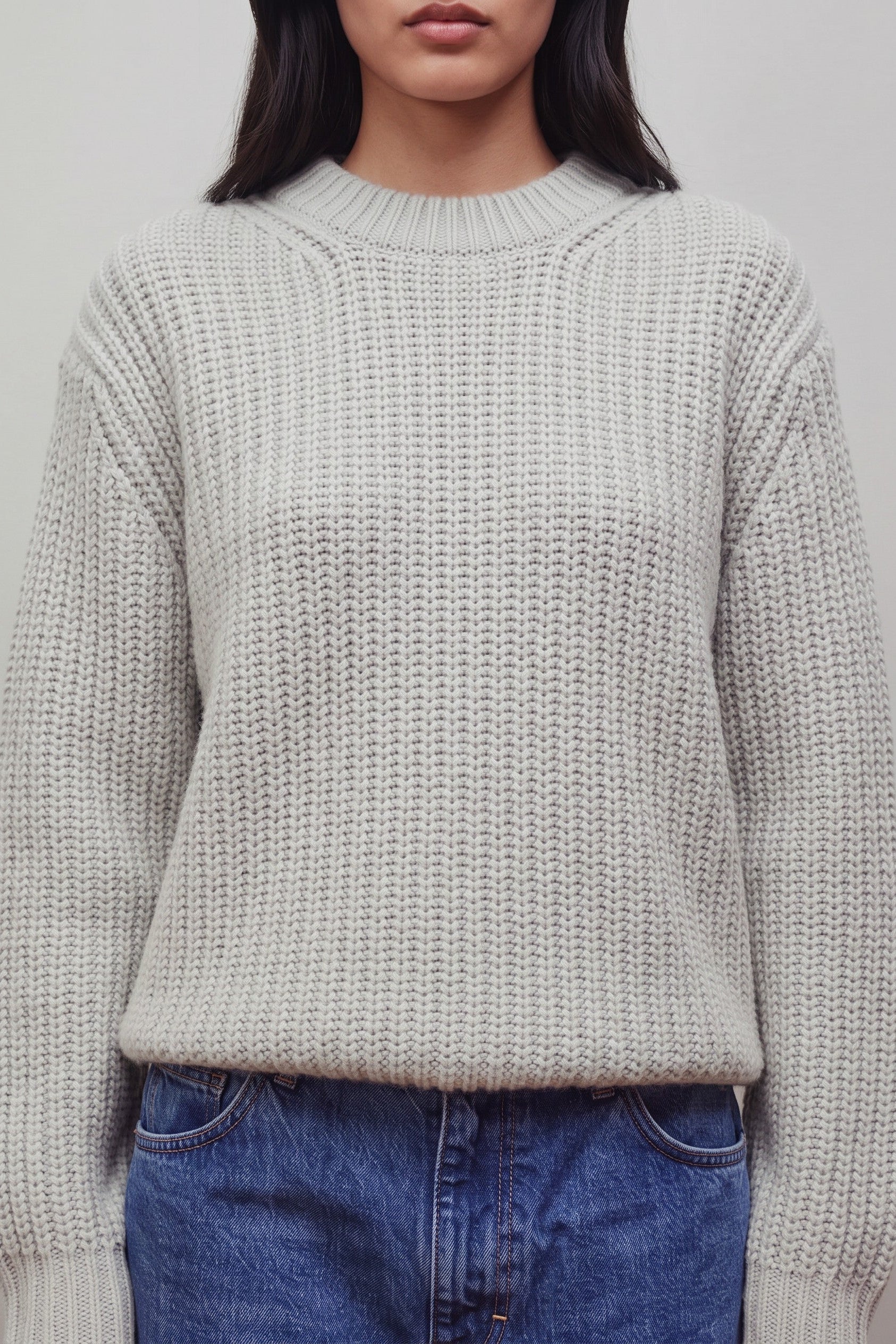 A close-up of a person wearing a chunky knit crewneck sweater in a light oatmeal gray color, paired with blue jeans against a plain light gray background.