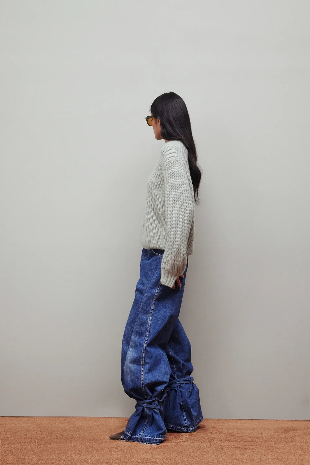 A woman stands in profile wearing a light gray ribbed knit sweater, blue wide-leg jeans with gathered ruffle details at the hems, and yellow-tinted sunglasses, set against a plain light gray wall and a brown wooden floor.