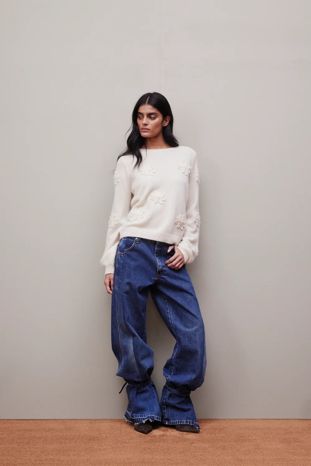 A woman stands against a plain light gray wall wearing a cream knit sweater with raised floral embroidery and wide-leg dark blue jeans.