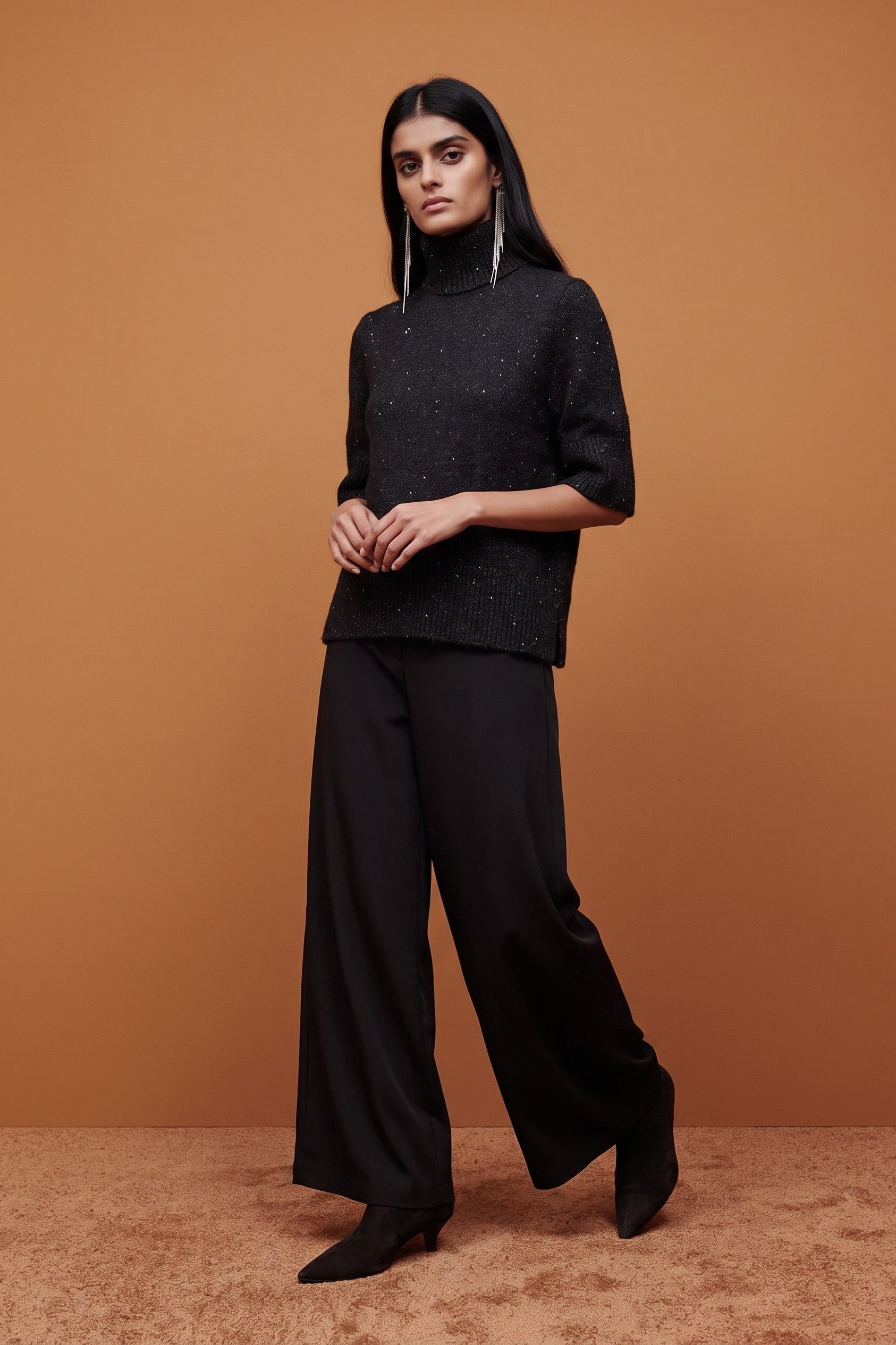 A model wearing a black textured turtleneck top, black wide-leg trousers, and black pointed-toe heels, standing against a solid warm brown studio backdrop.