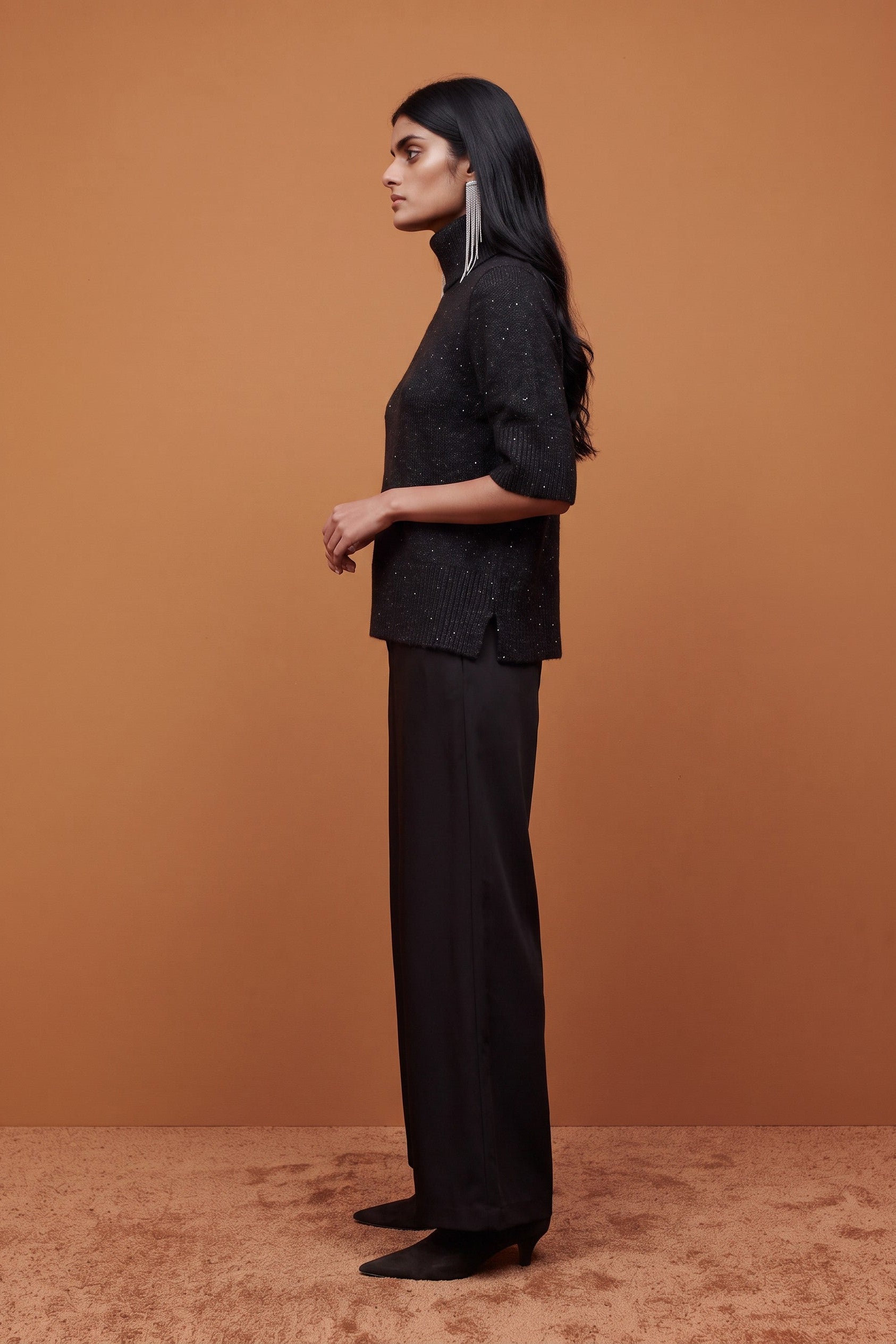 A woman stands in profile wearing a black textured short-sleeve top and black wide-leg trousers against a warm brown studio backdrop.