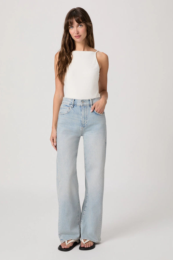 Woman wearing light wash barrel leg jeans with a white tank top and flip flops on a plain background.