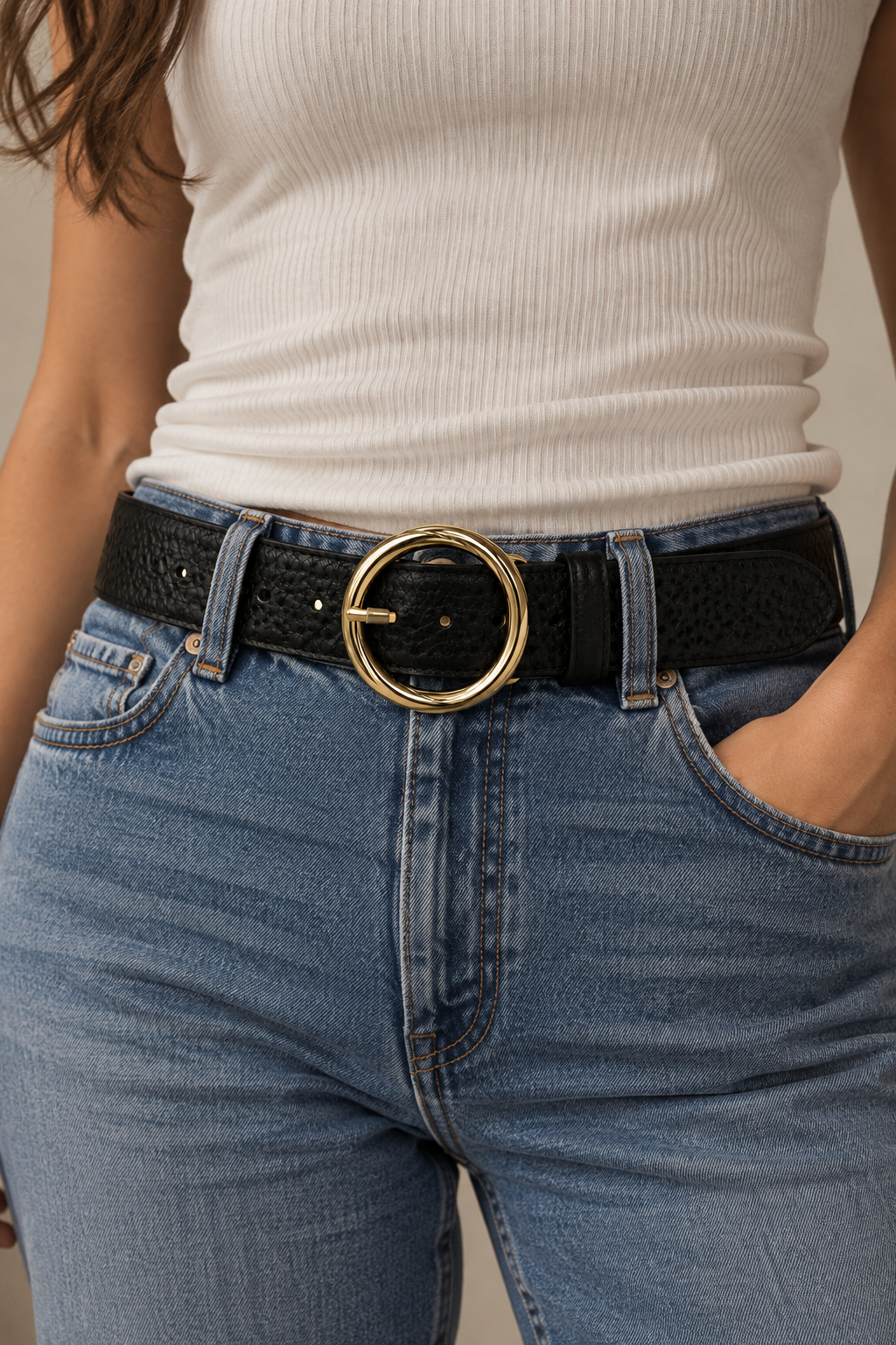 Close-up model view of a wide black textured leather belt with an oversized gold-tone circular buckle, styled with blue denim jeans and a cream ribbed top.