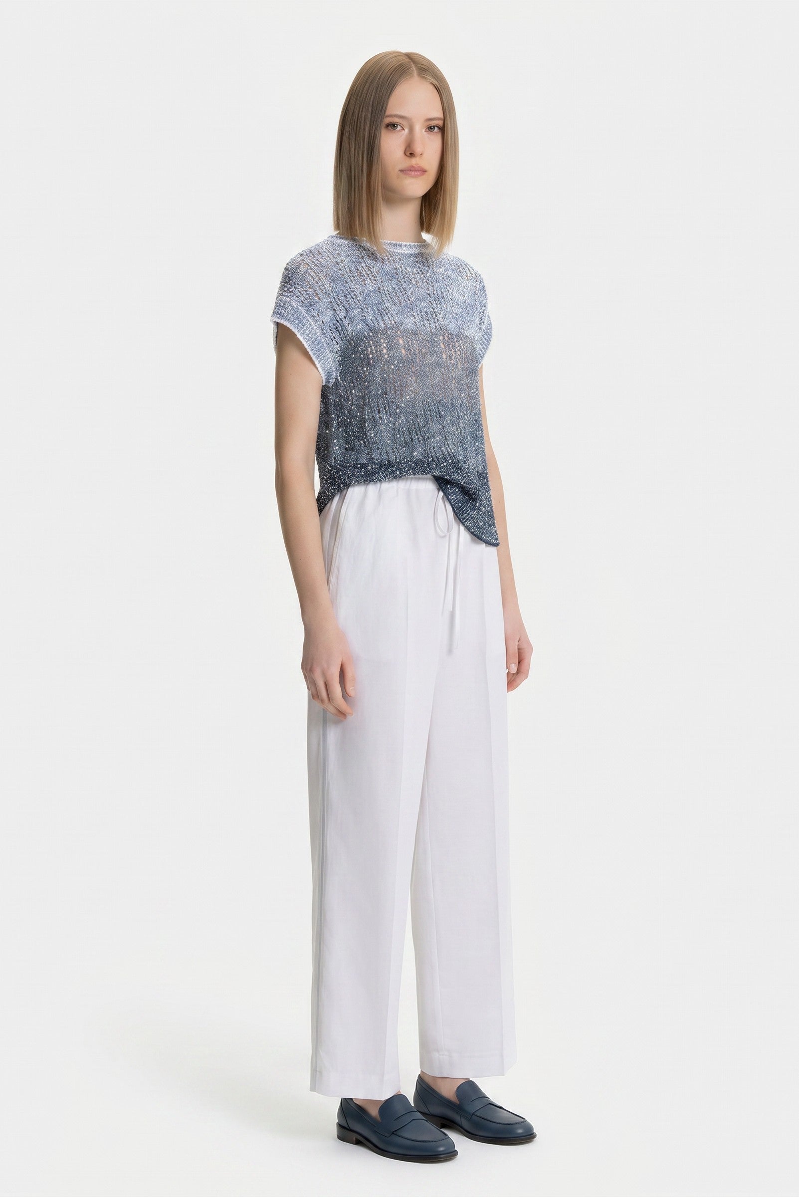 Woman wearing a patterned top and white pants on a white background.