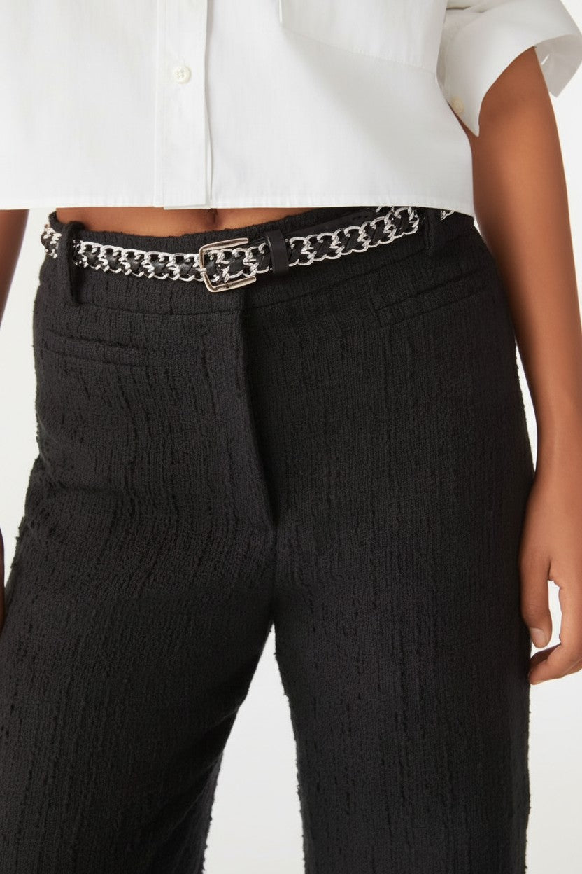 Close-up of a person wearing black textured trousers with a silver chain belt and a white cropped shirt, against a plain light background.