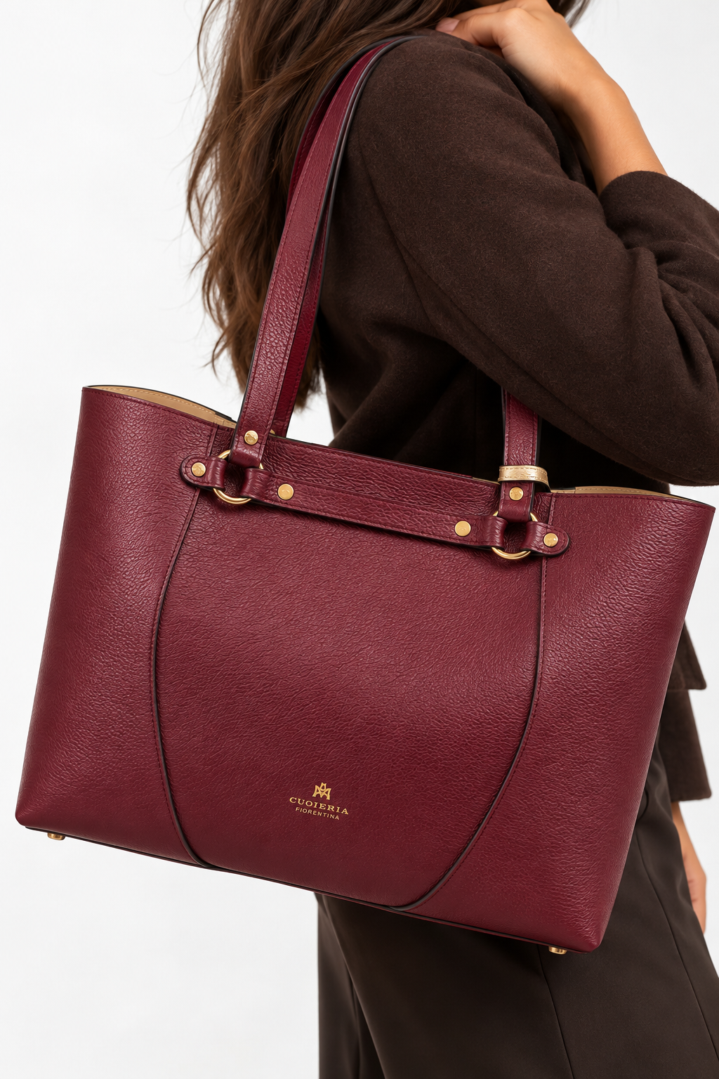 Close-up model shot of the Cuoieria Fiorentina Clara Shopping Bag in rich Bordeaux pebbled leather, featuring gold-tone hardware, double shoulder straps, and signature logo detailing, styled with an elegant chocolate brown outfit.
