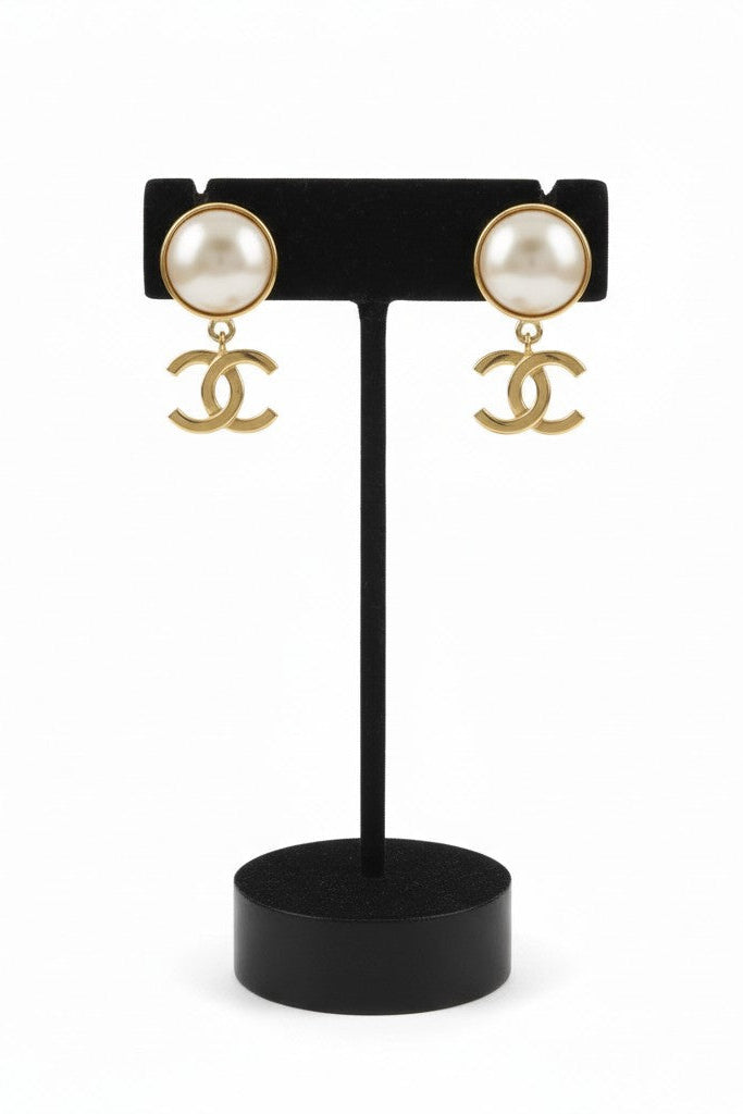 Pair of pearl earrings with gold CC logo on a black stand against a white background.