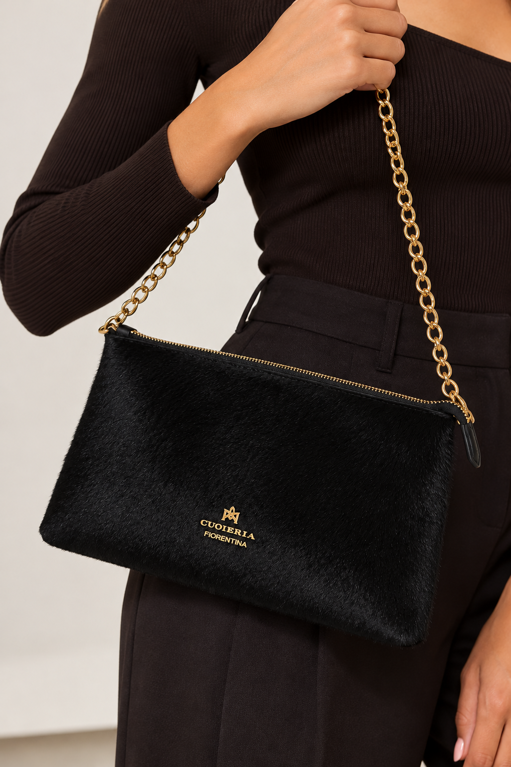 Close-up of a model holding the Cuoieria Fiorentina black calf hair pouch, featuring a gold-tone chain strap, zip-top closure, and signature gold logo, styled with an elegant black ensemble.