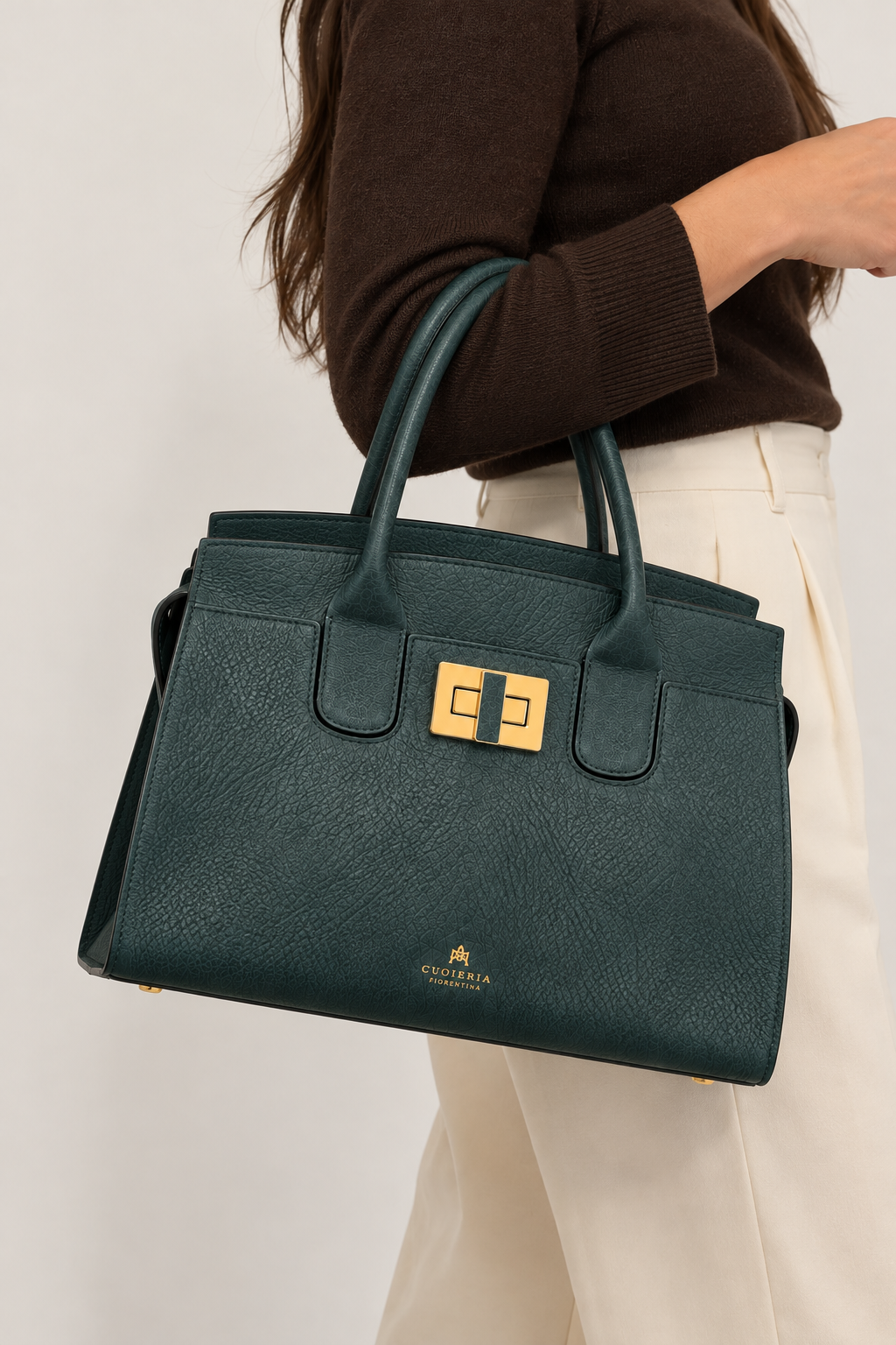 Model carrying the Cuoieria Fiorentina Bella Soft Medium Tote in deep ocean green pebbled leather with gold-tone hardware, styled with a chocolate brown sweater and ivory trousers against a clean neutral background.