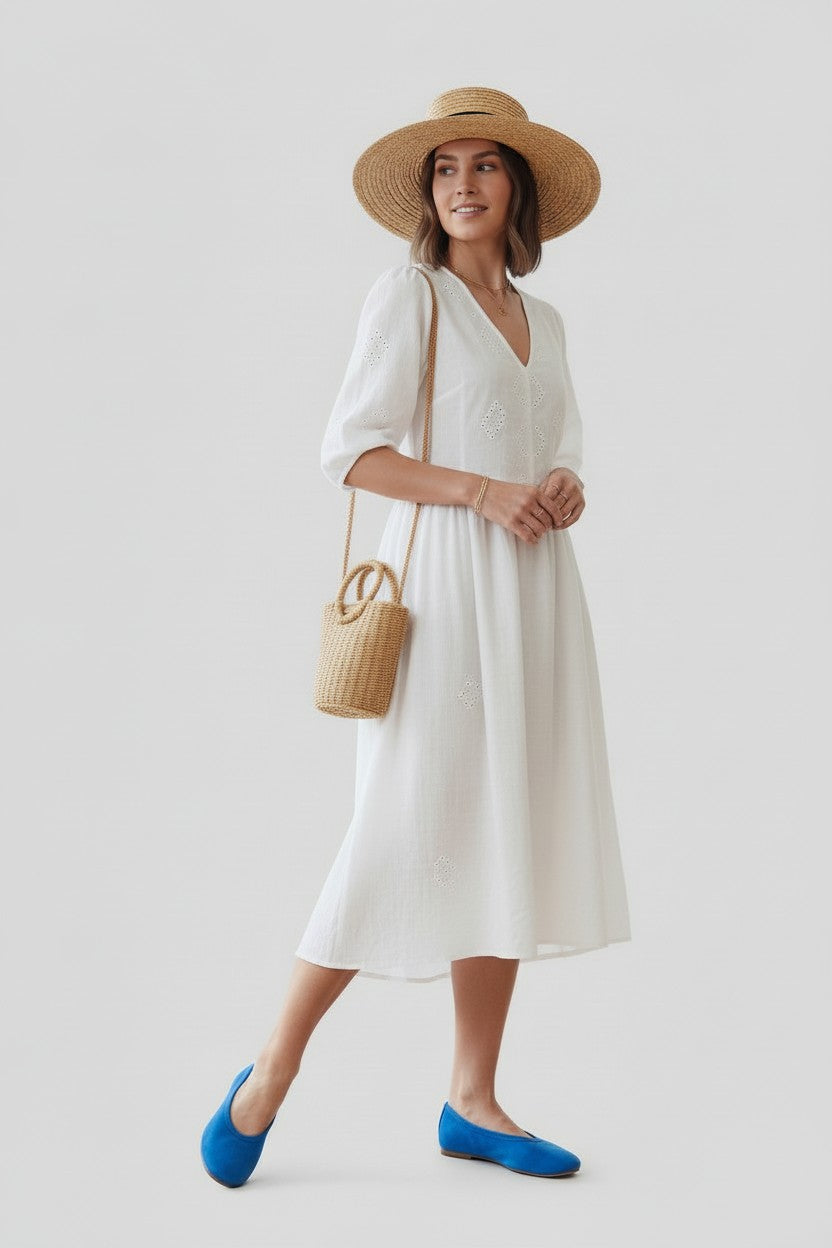 Woman wearing a white dress, straw hat, and a pair of blue flats on a plain background.