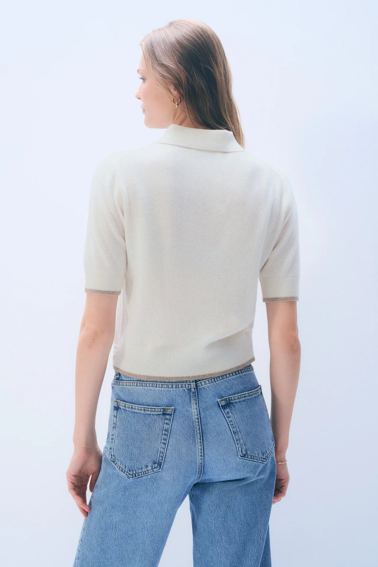 Back of a woman wearing a beige short-sleeve shirt and blue jeans against a light gray background.