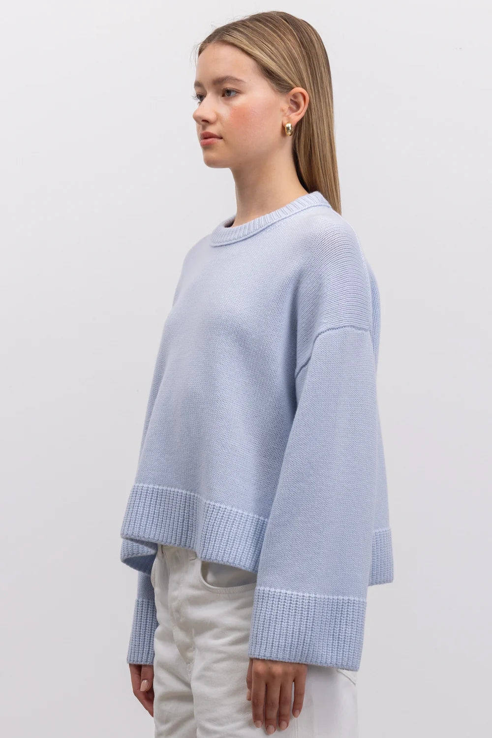 A woman wearing a light blue cropped knit sweater with ribbed cuffs and hem, paired with white pants, photographed against a plain light gray studio background.