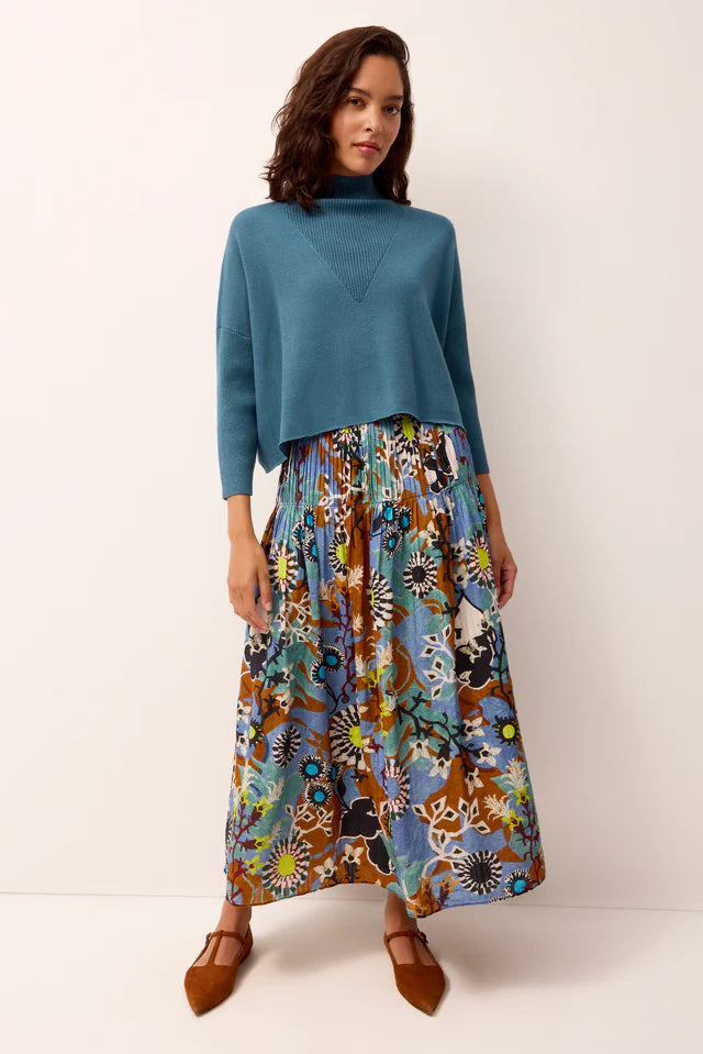 Woman wearing a teal sweater and a colorful floral skirt on a white background.