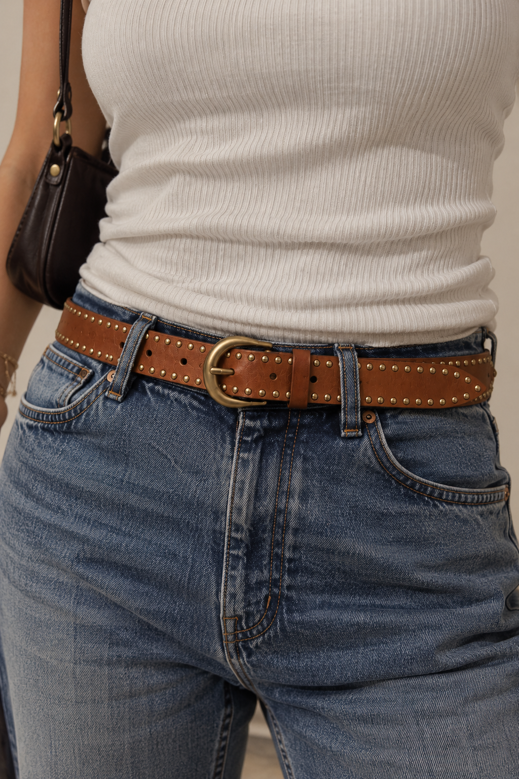 Close-up model view of a cognac brown leather belt with antique brass-tone studs and a rounded buckle, styled with blue denim jeans and a cream ribbed top.
